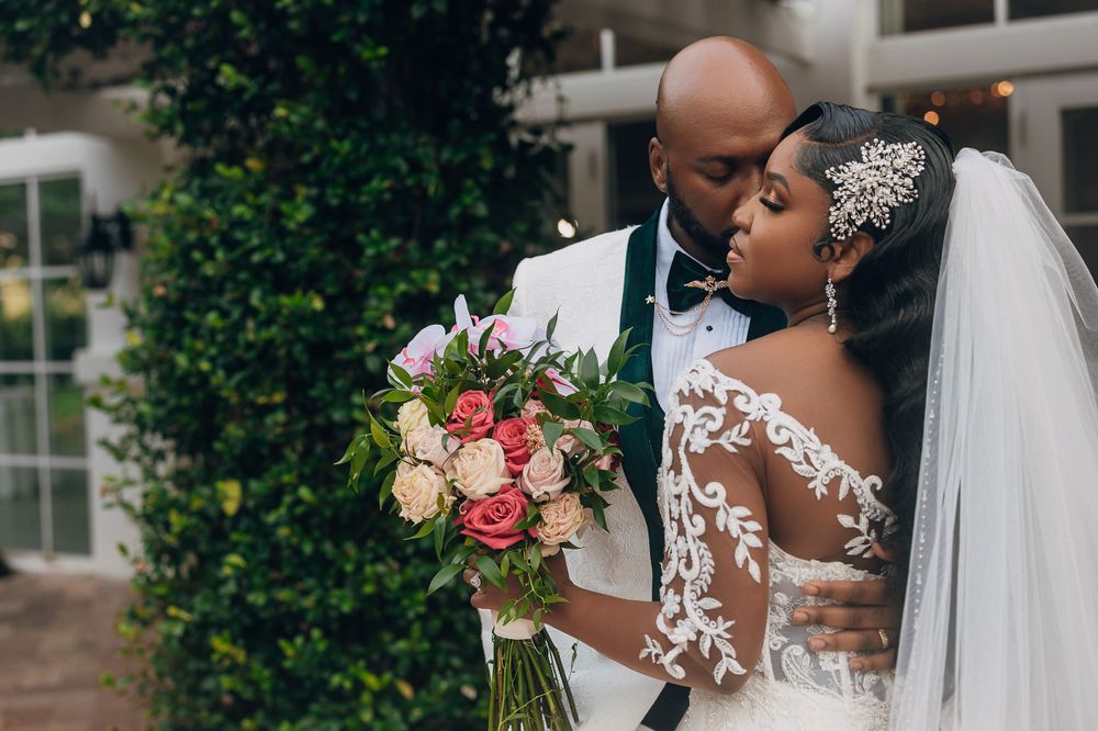 Groom in white tuxedo kisses bride wearing lace long-sleeve wedding dress while holding floral bouquet.