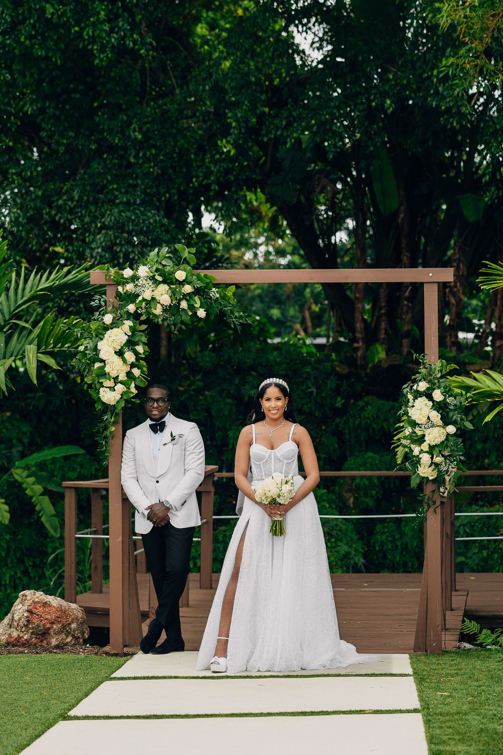 Elegant couple poses under floral wooden arch at outdoor wedding ceremony surrounded by lush greenery.