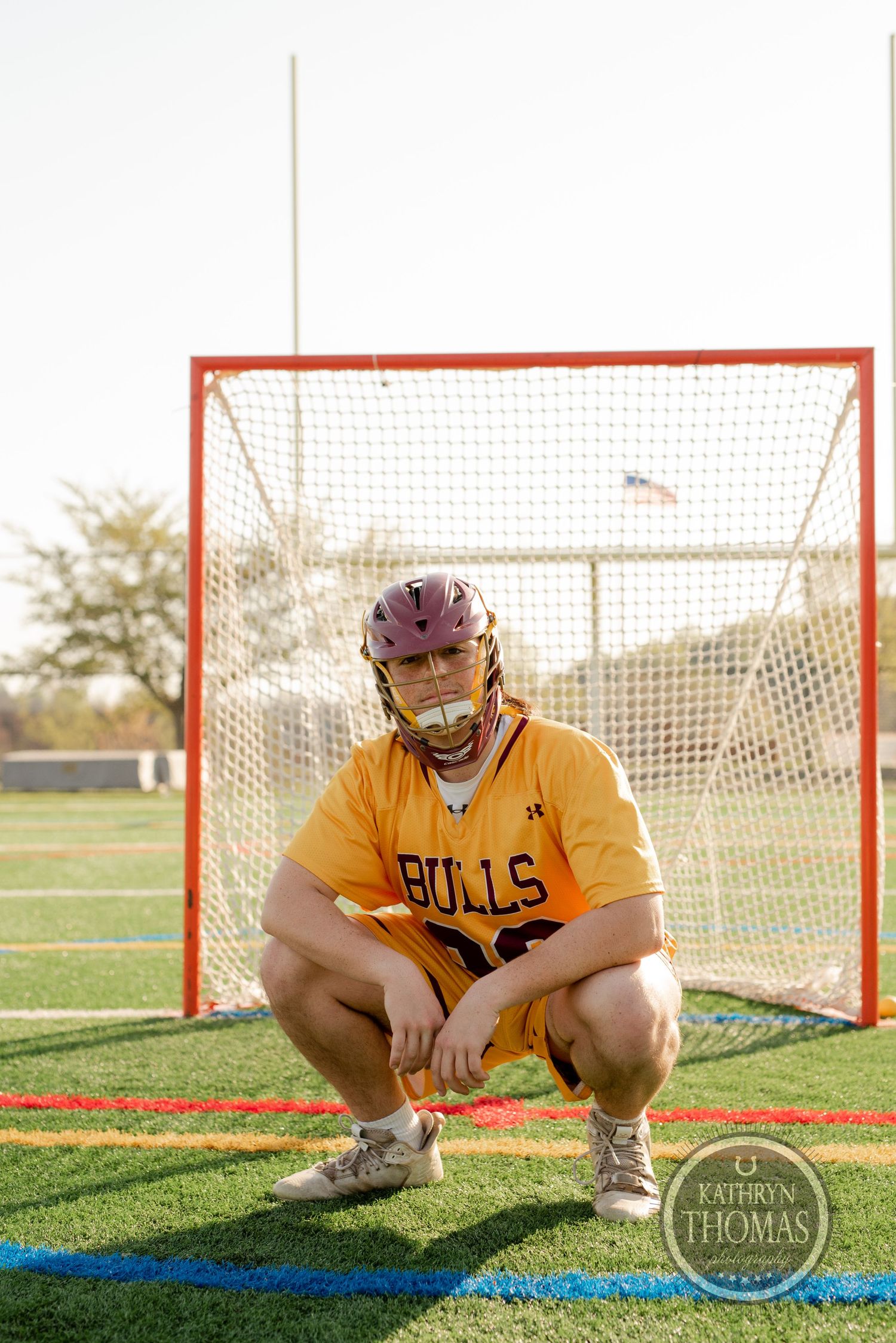 Lacrosse player in Bulls uniform crouching by goal net wearing purple helmet.