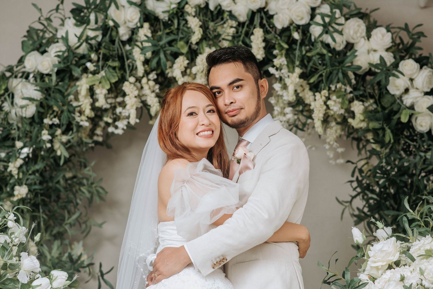 A wedding couple embraces under a lush floral arch with white roses and greenery.