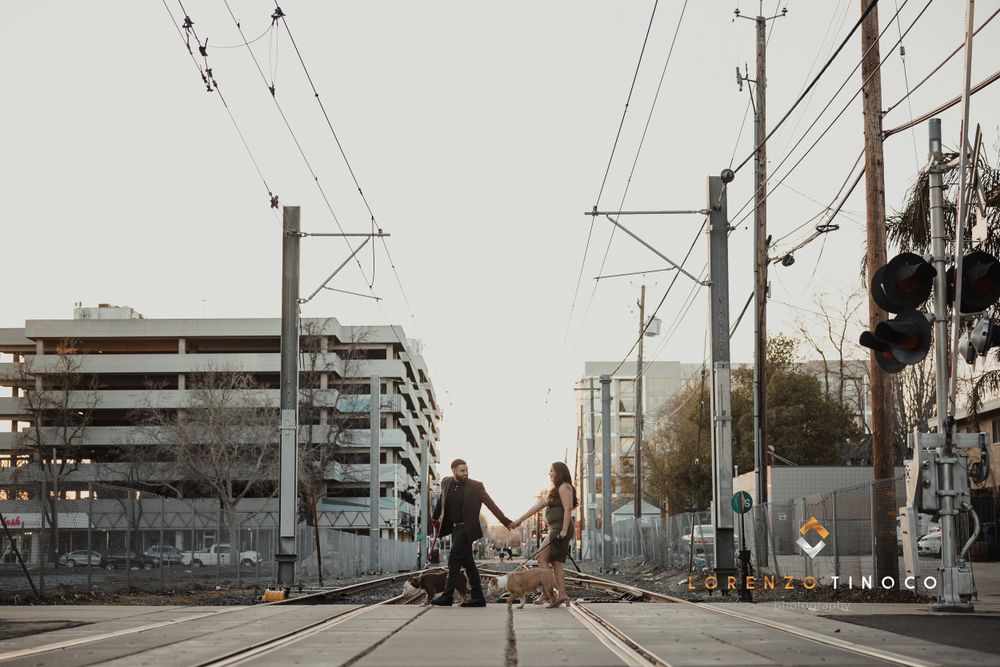 A couple walks hand in hand down railroad tracks in an urban setting with buildings and power lines in the background.