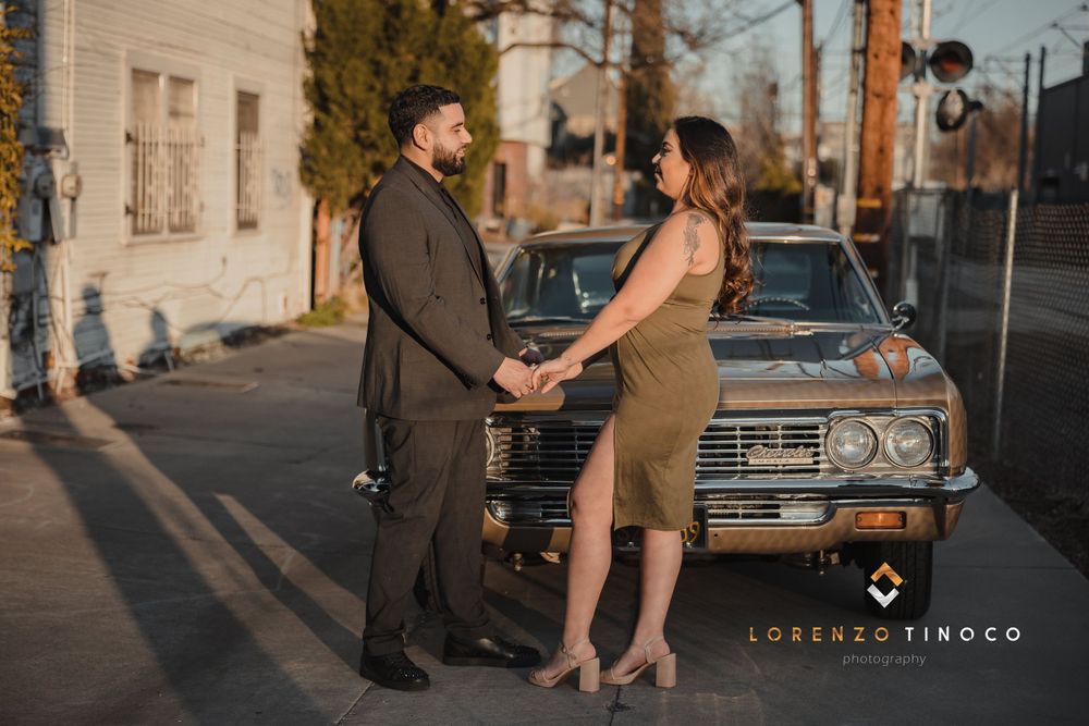 Romantic couple poses beside classic car during urban sunset photoshoot.