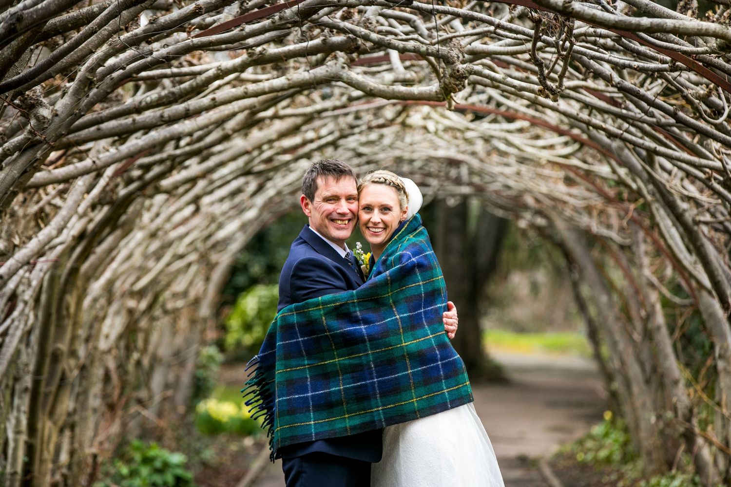 Newlyweds wrapped in a plaid blanket share a laugh under a rustic branch tunnel.