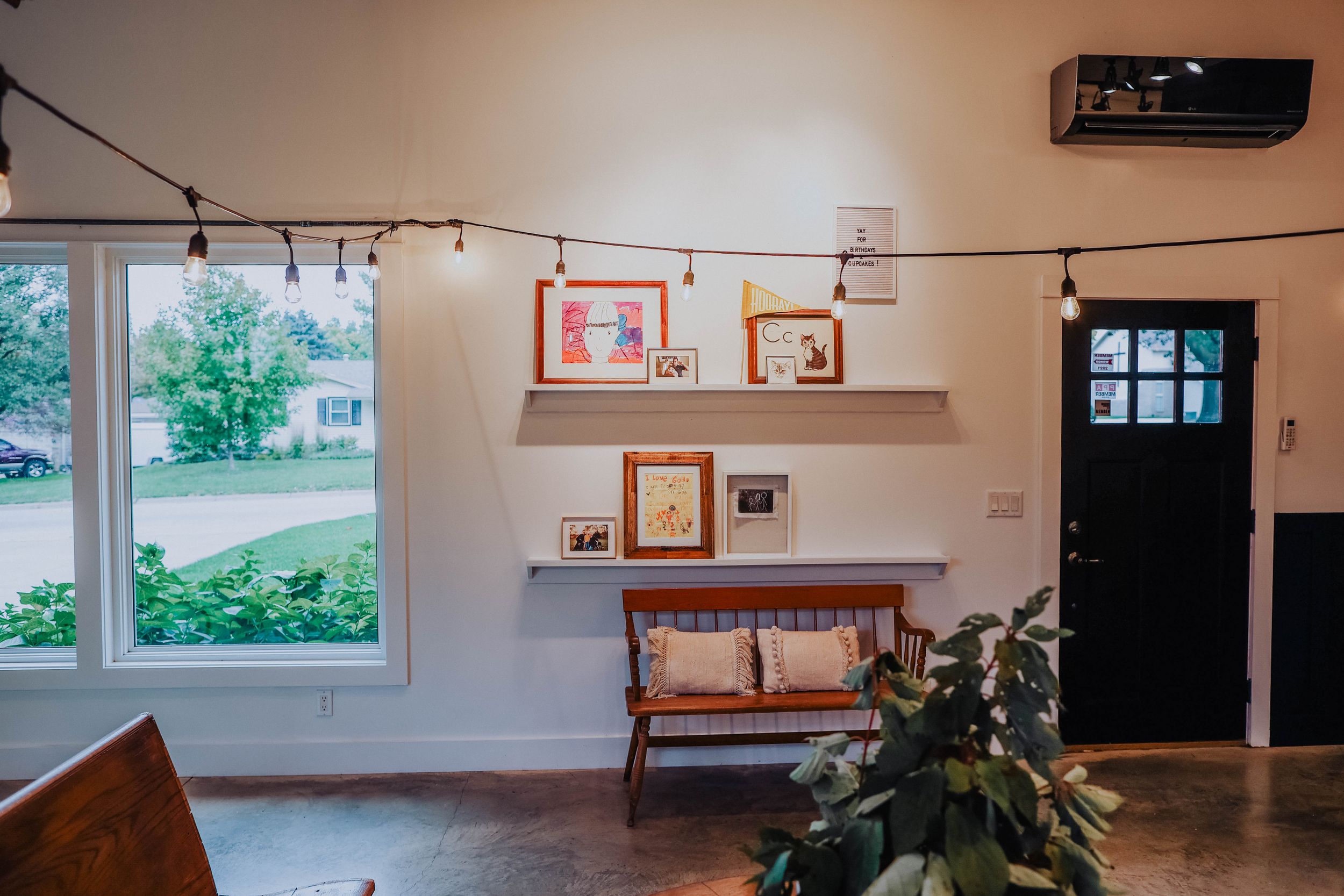 Bright interior space with large windows, string lights, and wooden furniture against white walls.