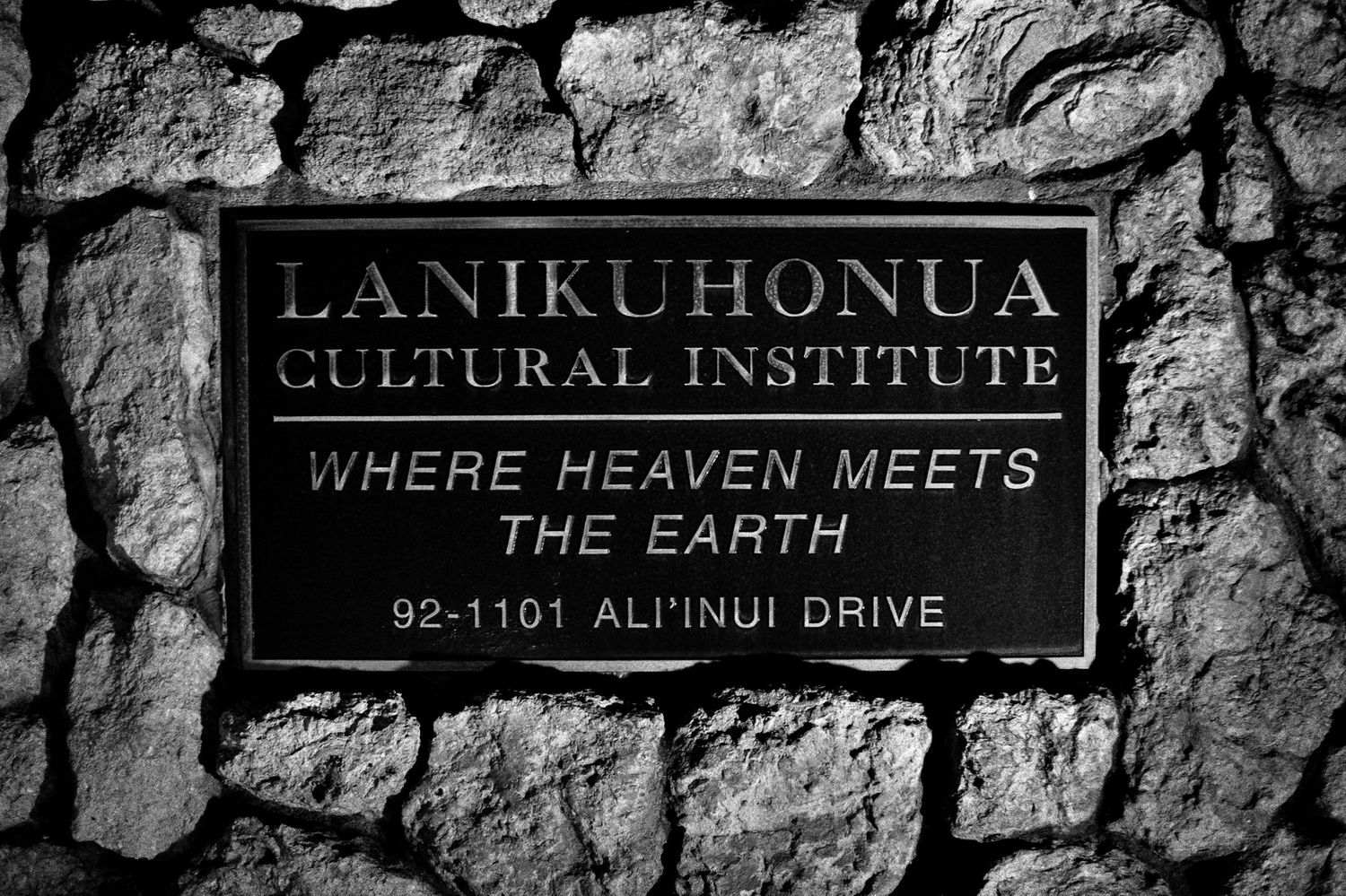 Black and white sign for Lanikuhonua Cultural Institute reading Where Heaven Meets The Earth on stone wall.