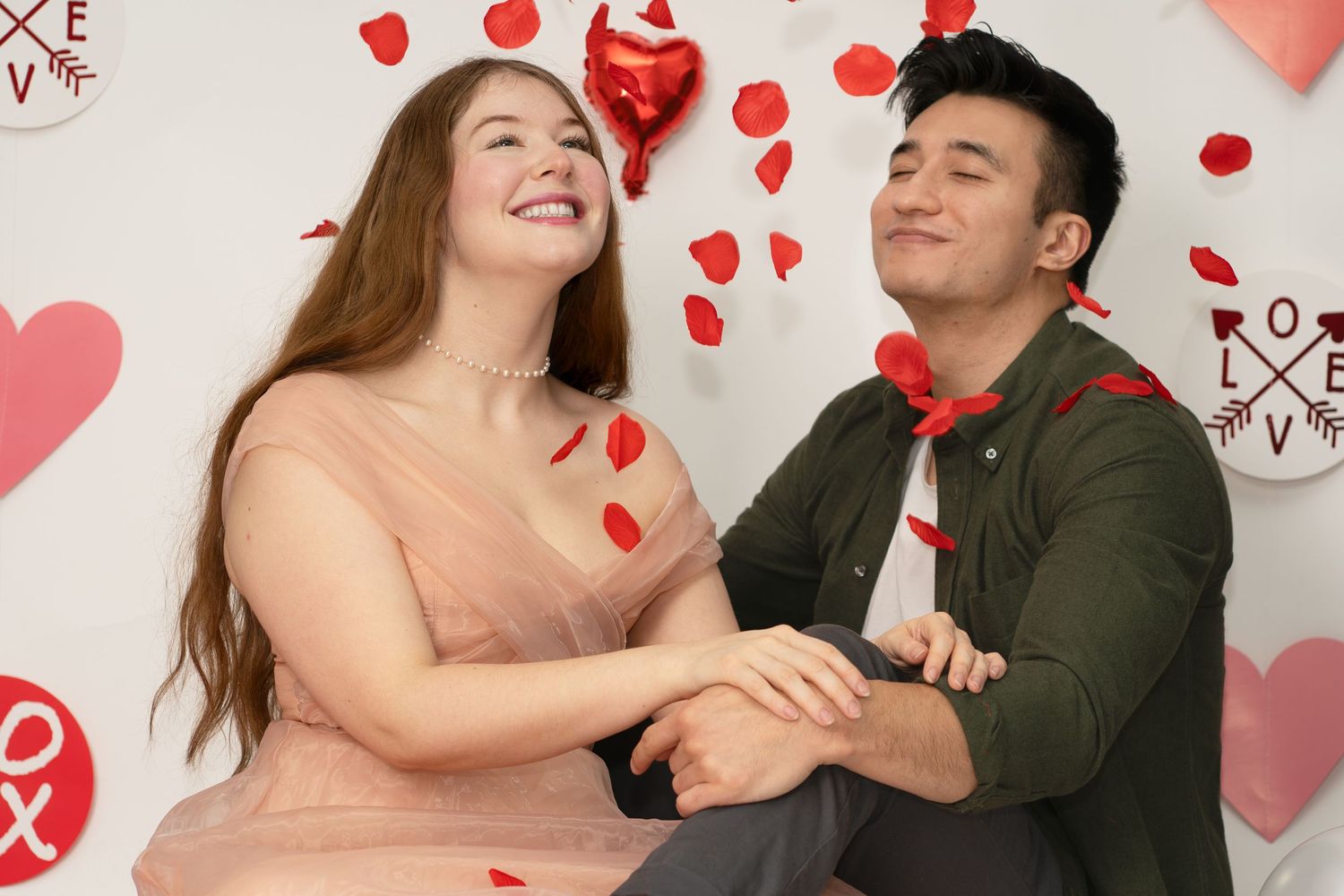 Romantic couple poses playfully with red heart decorations and balloons against white backdrop.