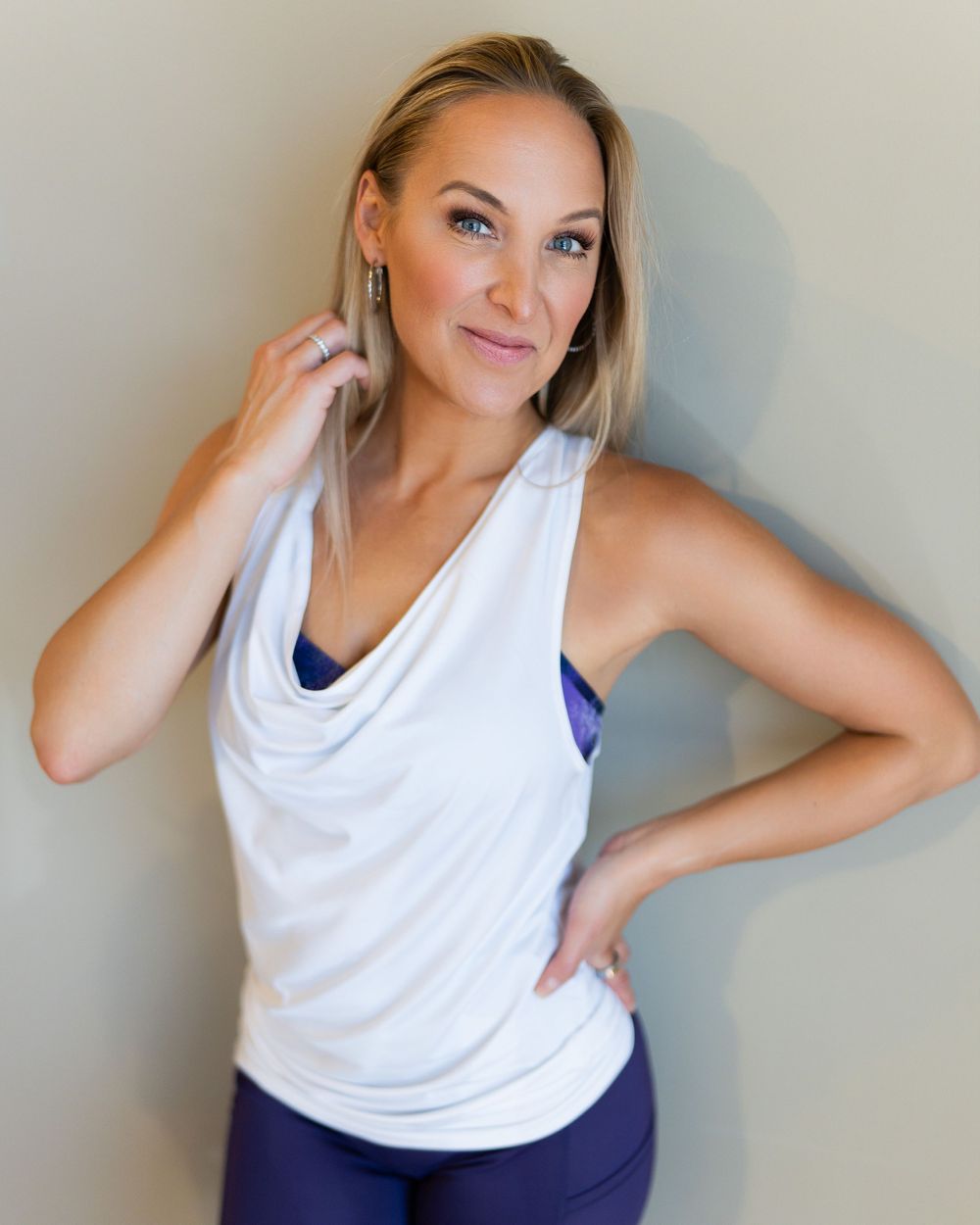 A model in a white cowl neck tank top and navy leggings poses against a neutral wall in Handful Activewear. Brand photography.