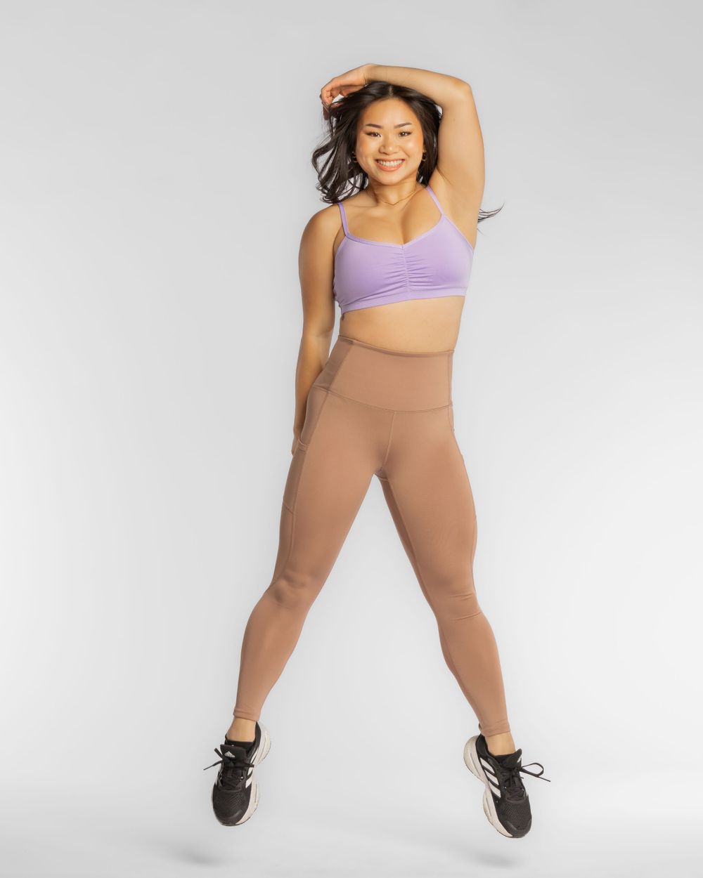 Athletic woman in purple sports bra and tan leggings jumping energetically against light gray background. Brand photography for ecommerce.