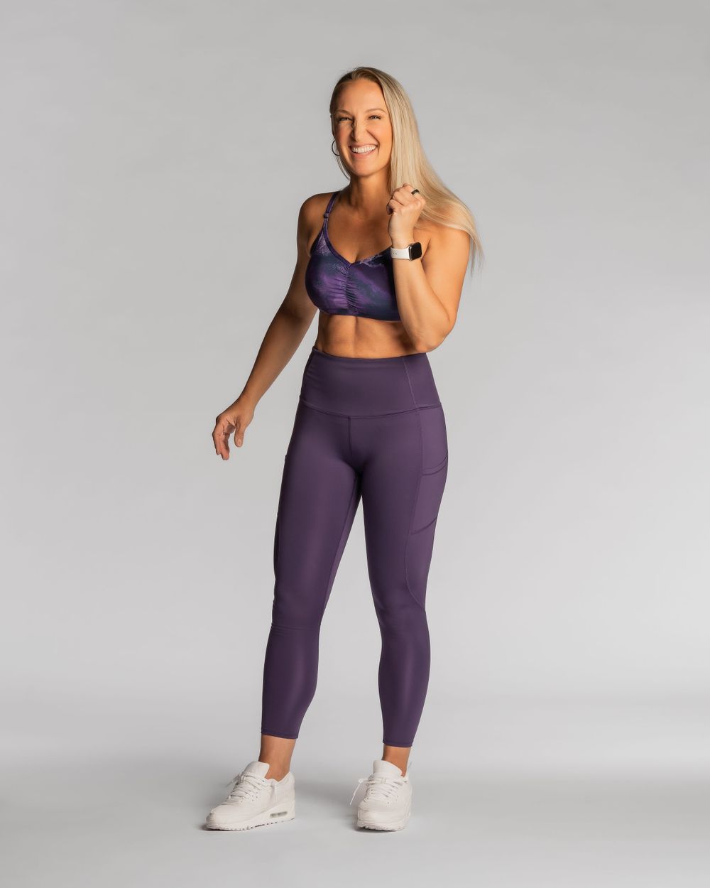 Athletic woman in Handful purple workout set and white sneakers poses against gray background. Brand photography.