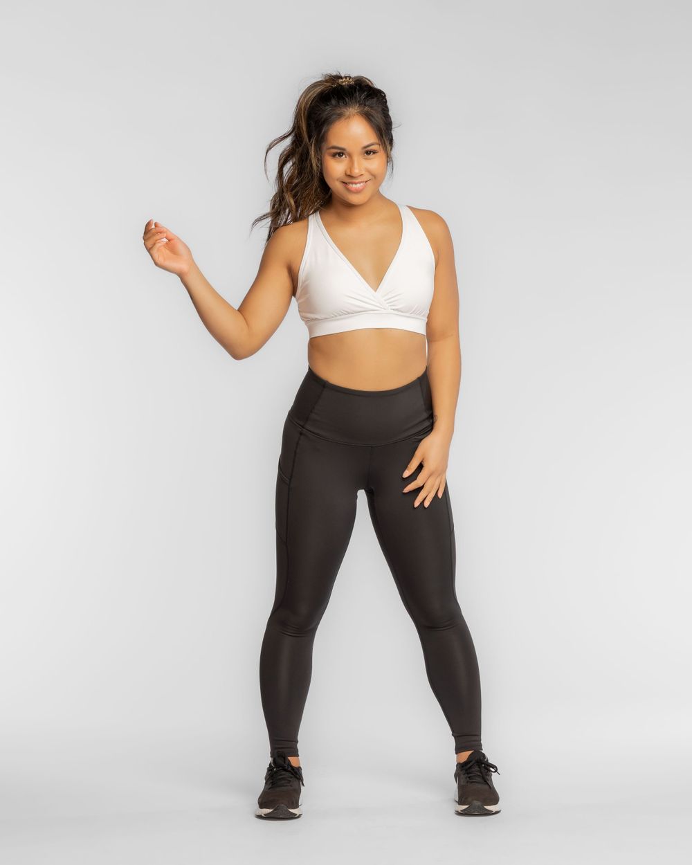 Athletic figure in white sports bra and black leggings poses against gray background in fitness apparel. Brand photography for women's wear.