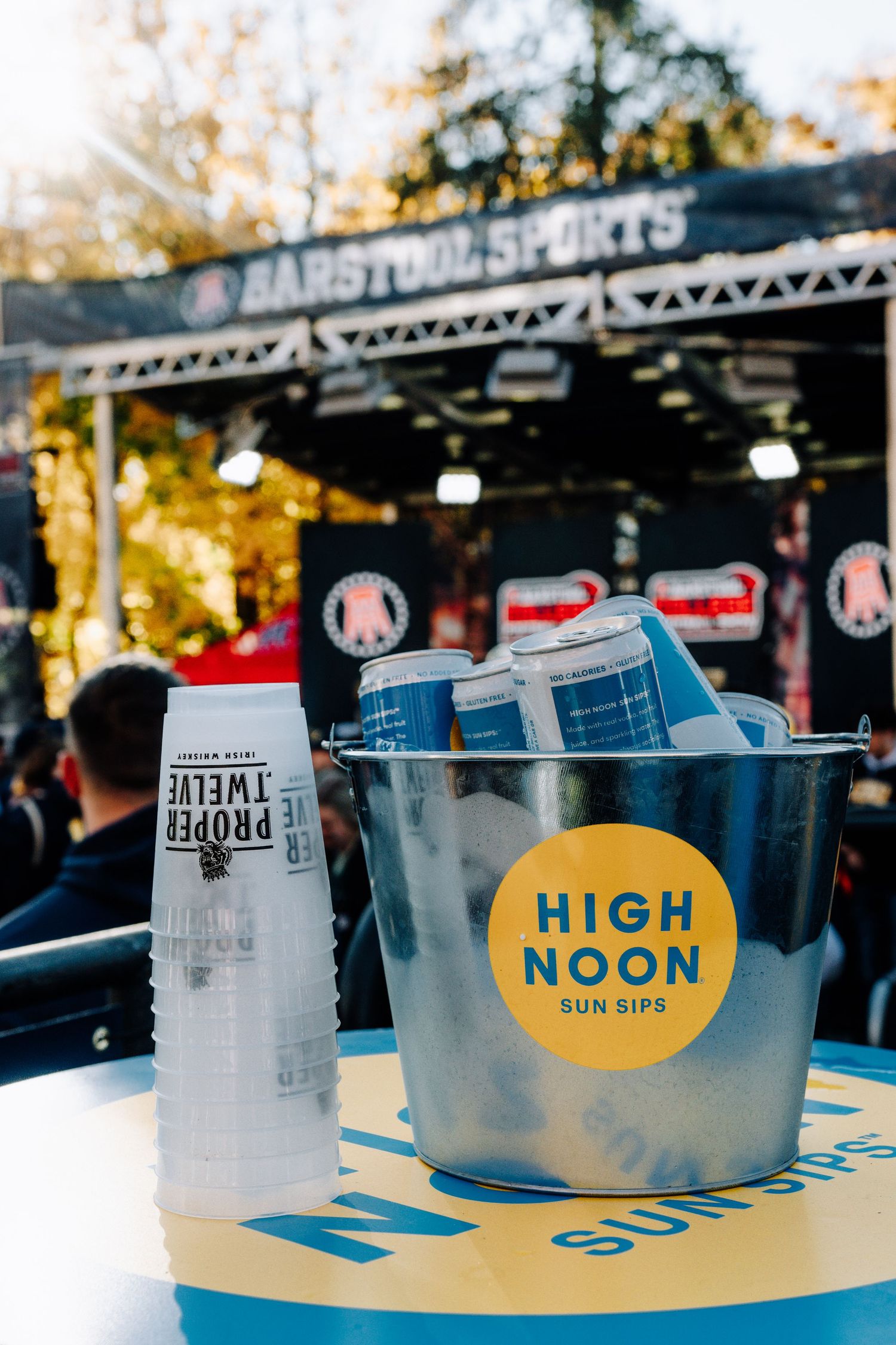 High Noon hard seltzer drinks in an ice bucket at an outdoor event with Barstool Sports branding.