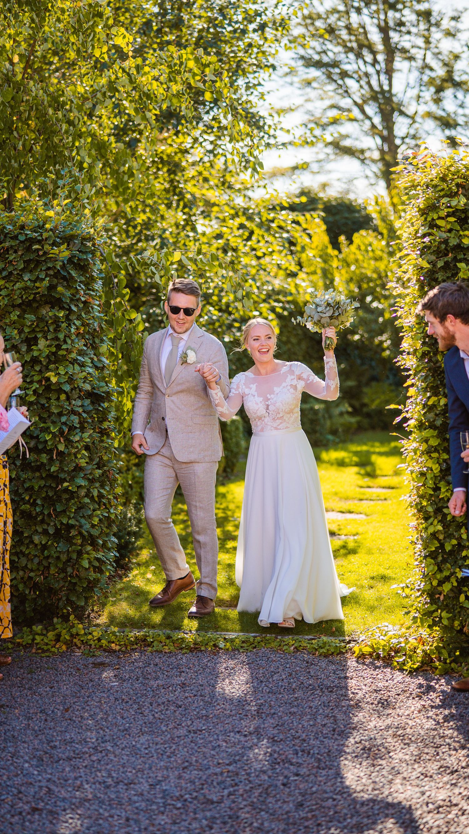 Happy newlywed couple walking through garden arch, bride holding bouquet aloft in white lace gown and groom in tan suit.