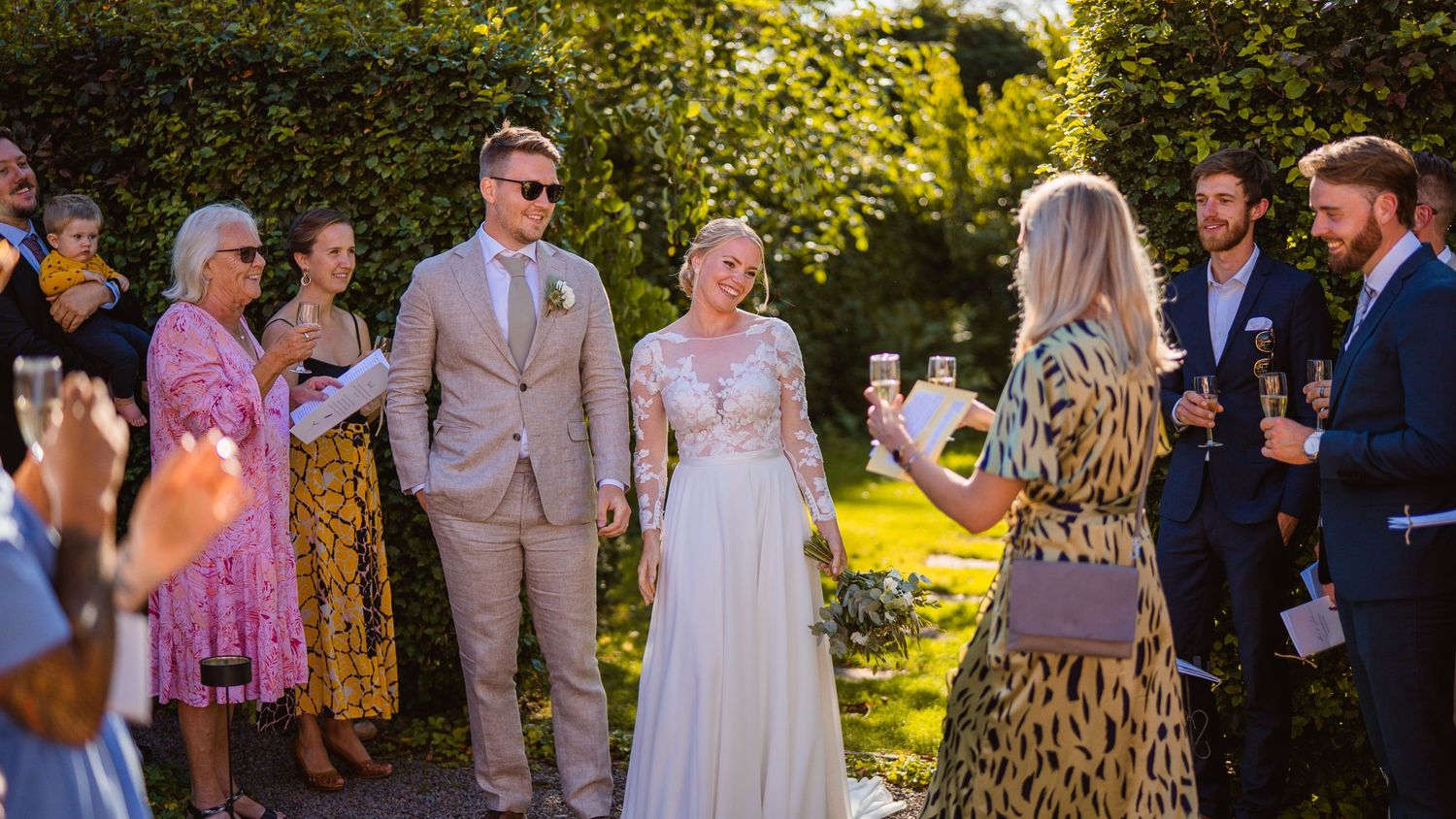 Wedding guests raising glasses to toast smiling newlywed couple in sunny garden setting.