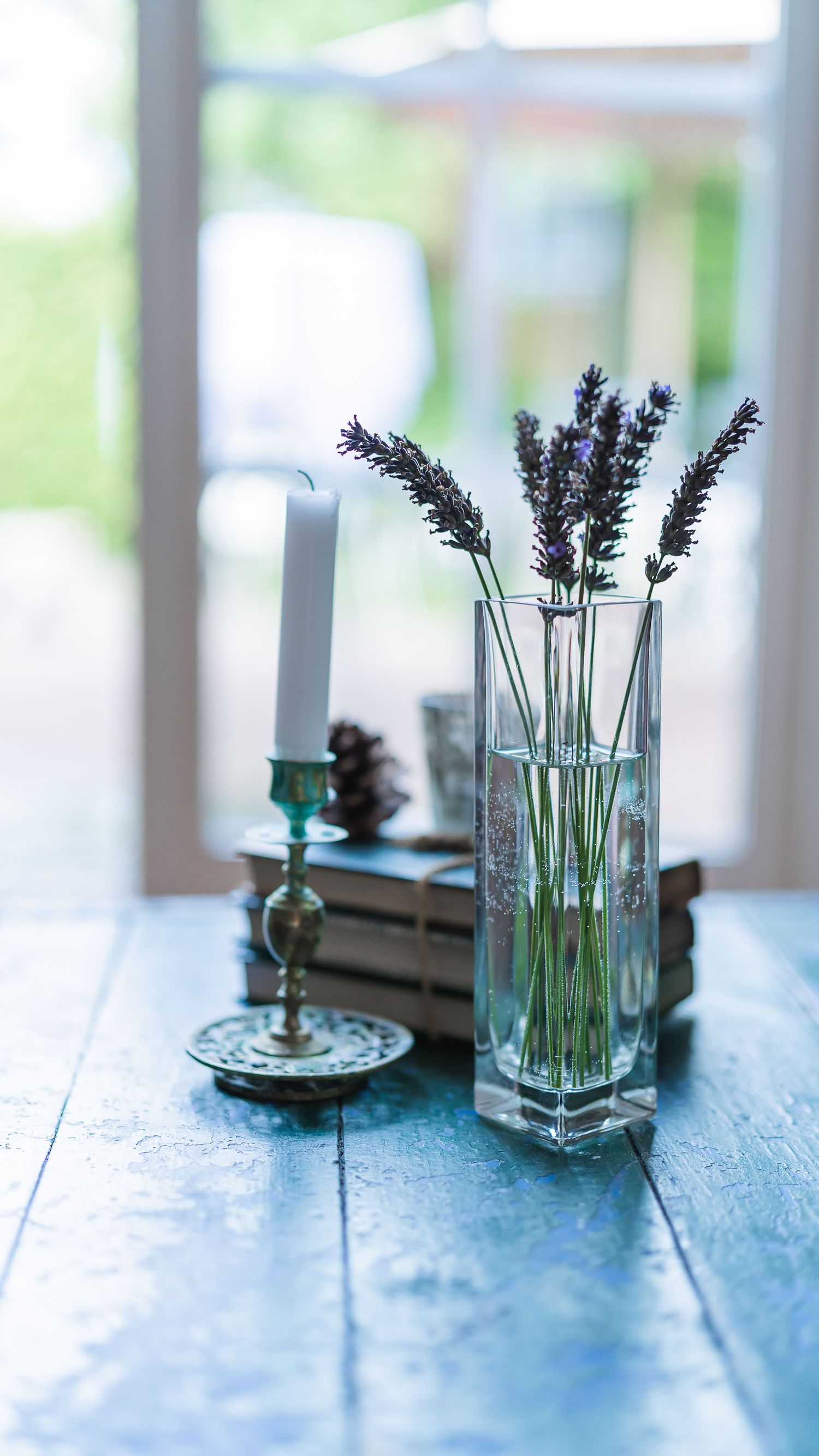 Glass vase with lavender stems beside a candle and books on a blue wooden table near a window.