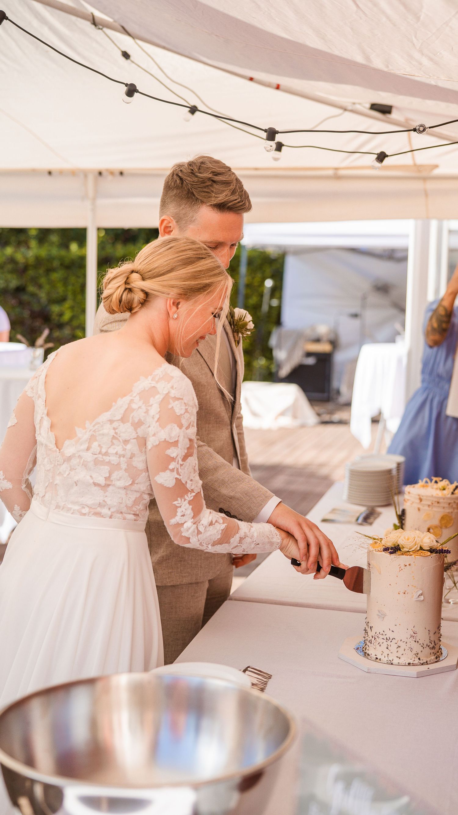 Bride and groom cutting elegant white wedding cake decorated with roses under a marquee tent.