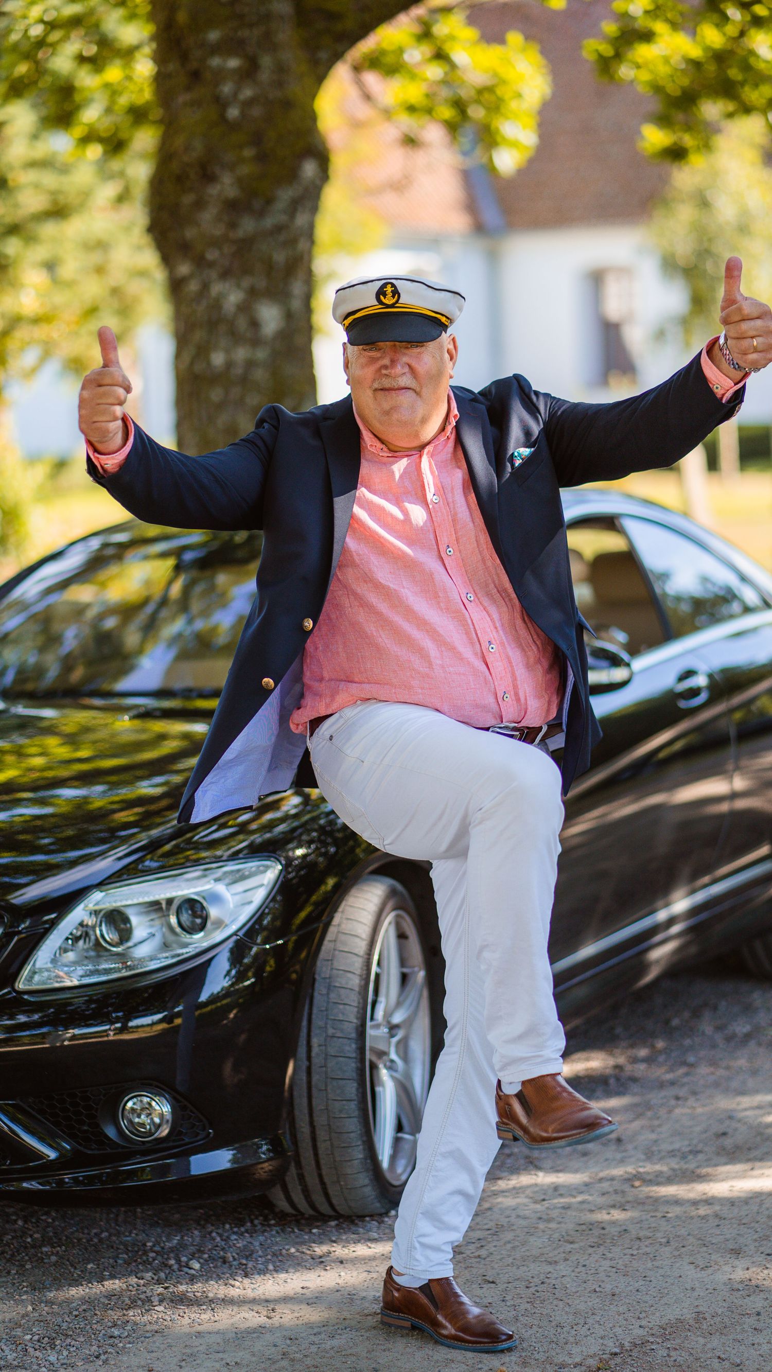 Cheerful man in captain hat and navy blazer giving thumbs up while leaning against a black Mercedes car outdoors.