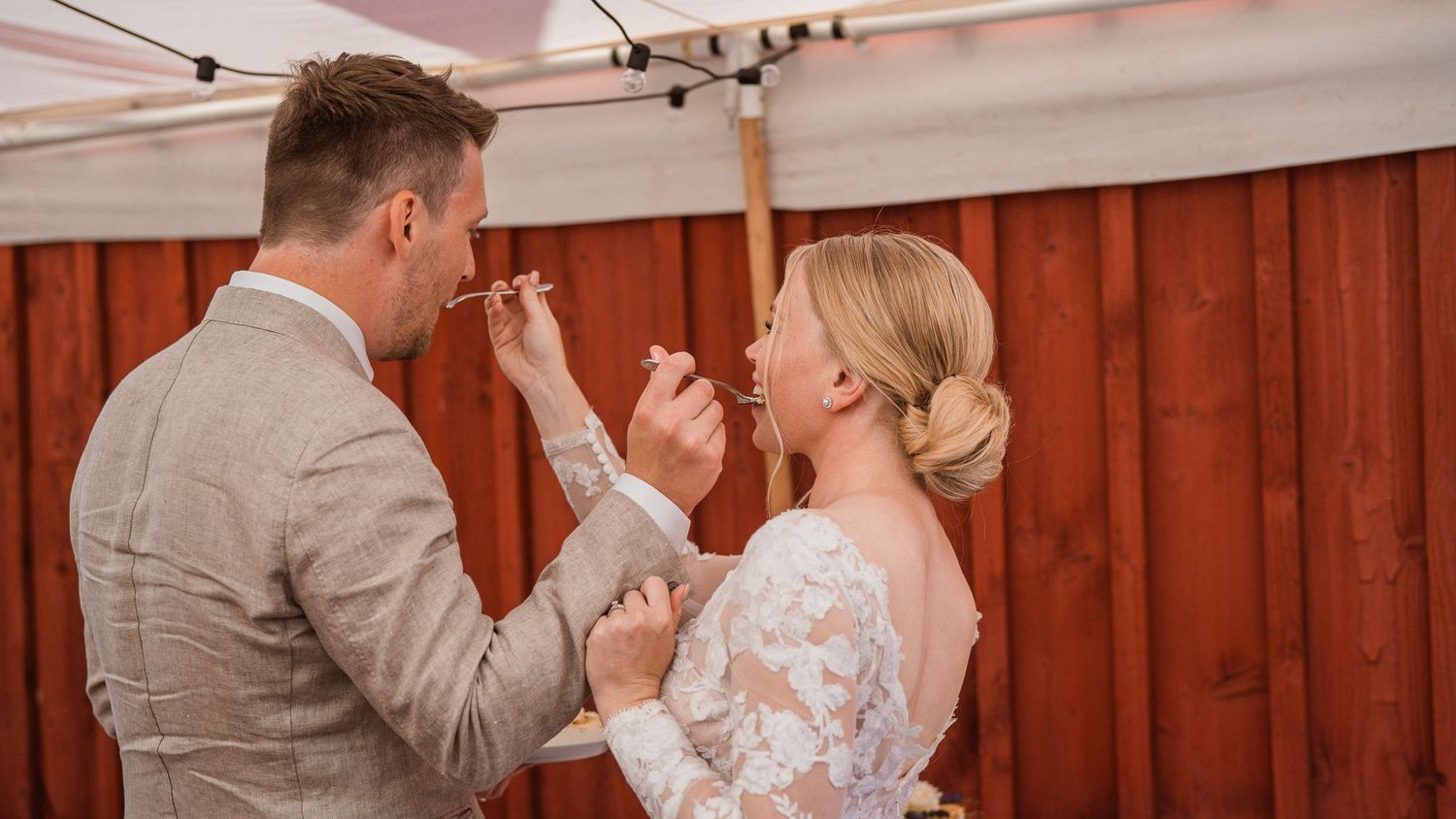 Newlyweds feeding each other wedding cake and laughing together in front of a red wooden wall.