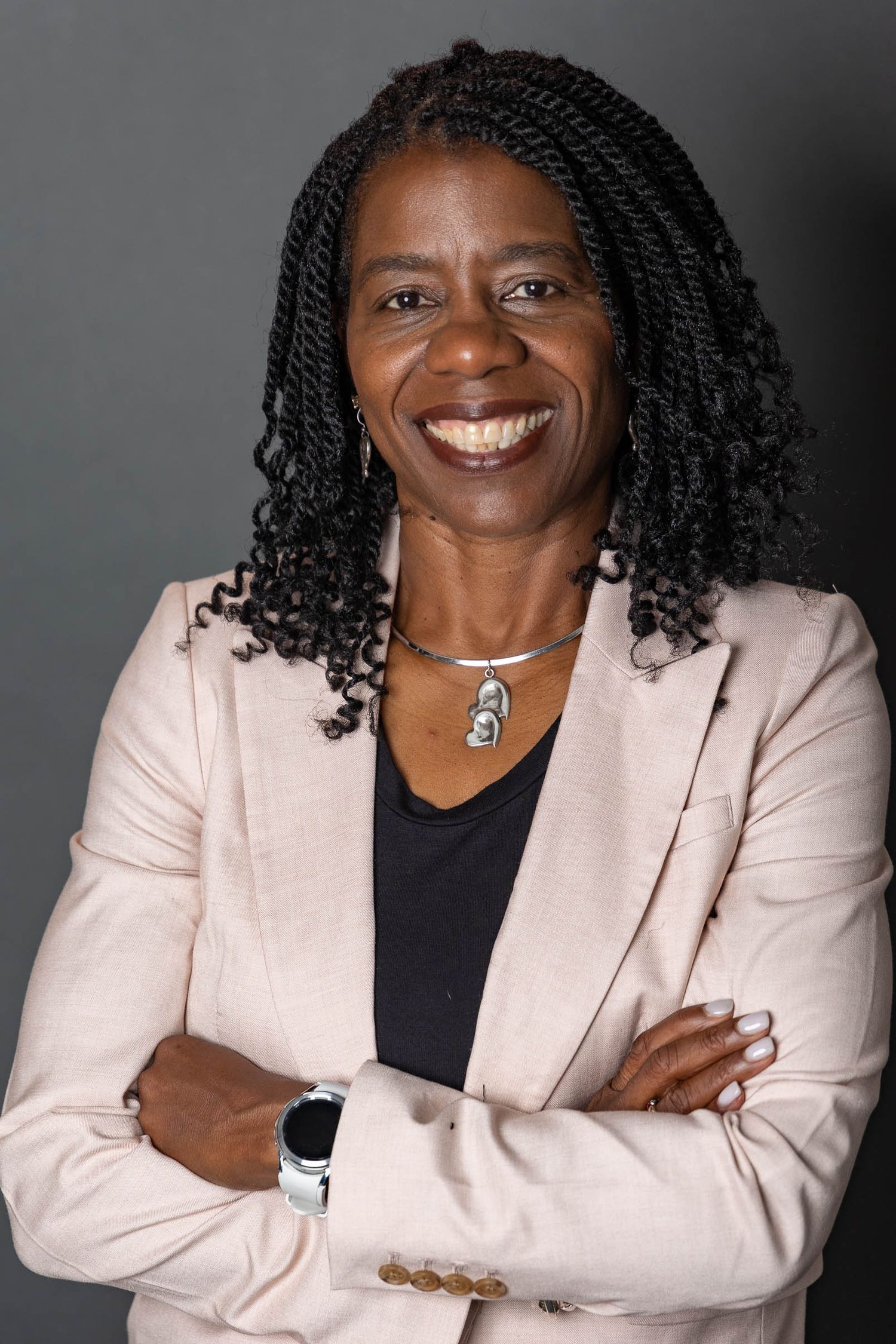 Professional portrait against gray background featuring pink blazer, black top and pendant necklace.