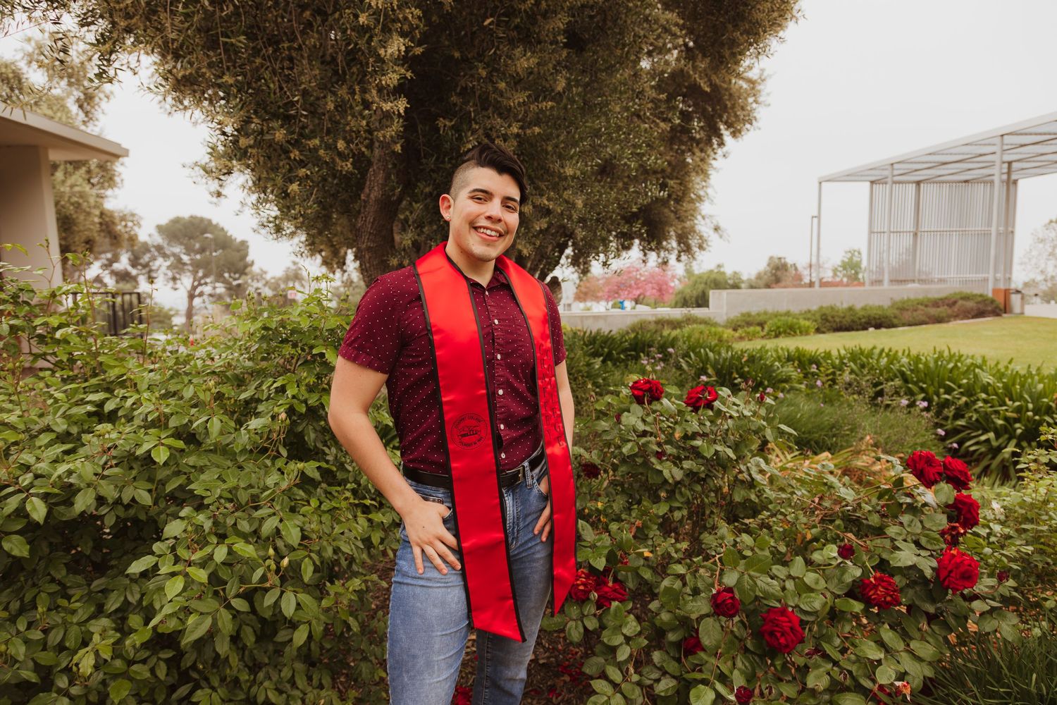 Graduate in red stole poses in garden with blooming red roses and trees.
