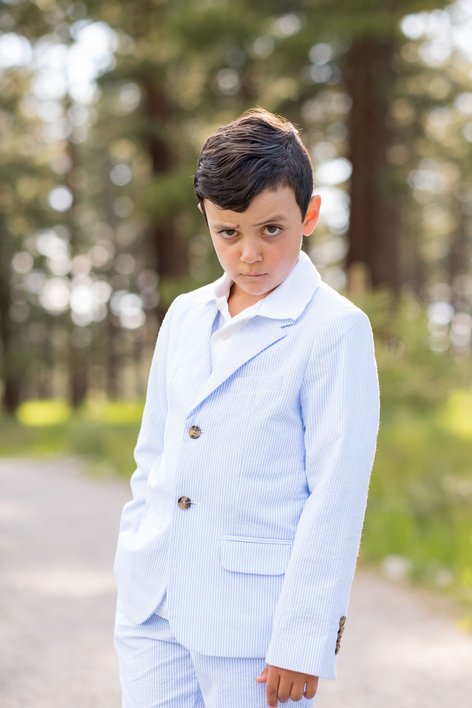 A child in a light blue seersucker suit stands outdoors on a path with trees in the background.