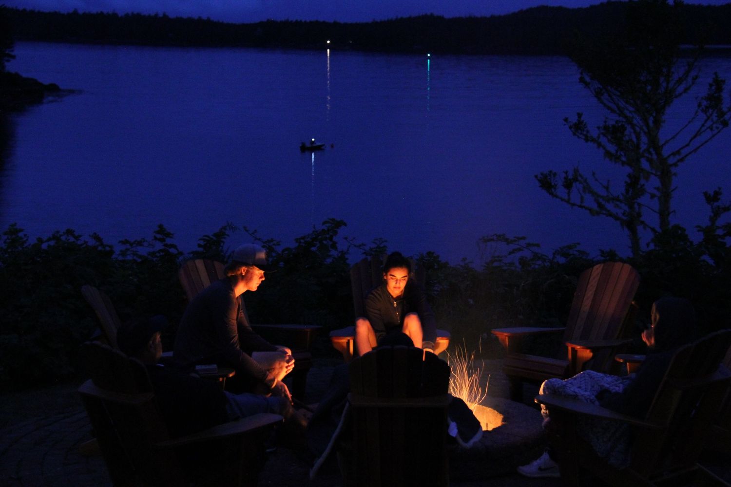 Group gathered around a campfire at night by a lake with boats in the distance.