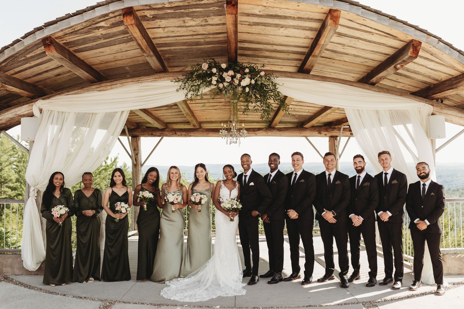 Wedding party poses under a wooden pavilion decorated with white draping and florals.