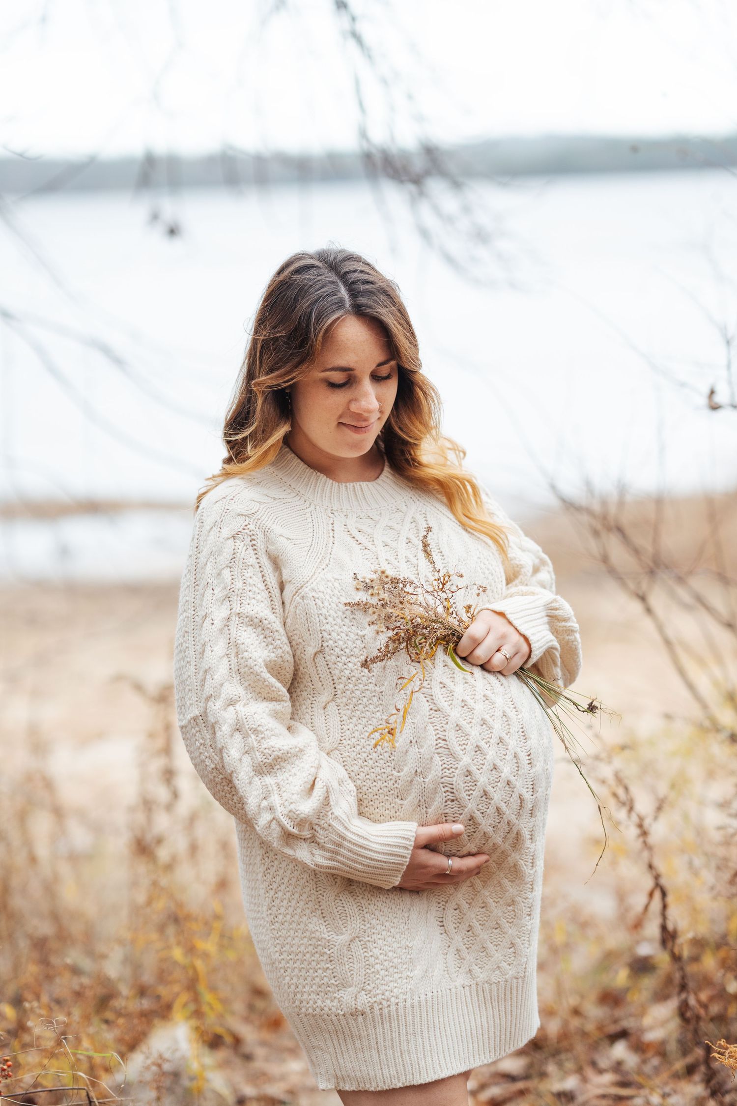 Woman in cream sweater poses with dried flowers in rustic winter maternity photos.
