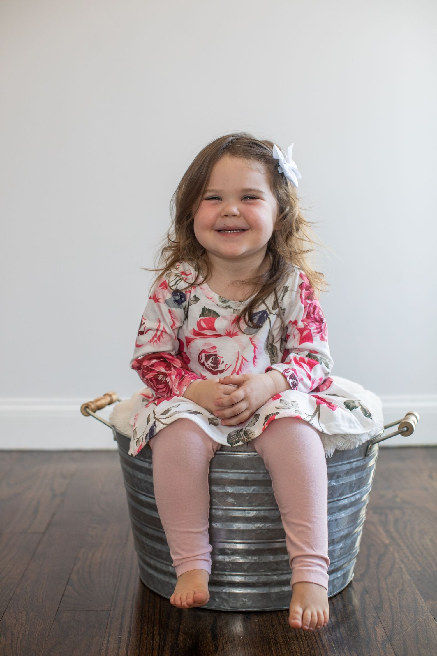 A young child in floral clothing sits cheerfully on a metal bucket against a white wall.
