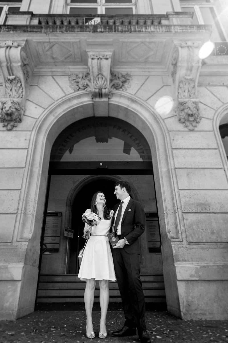 Black and white wedding portrait under ornate stone archway in classic architectural setting.