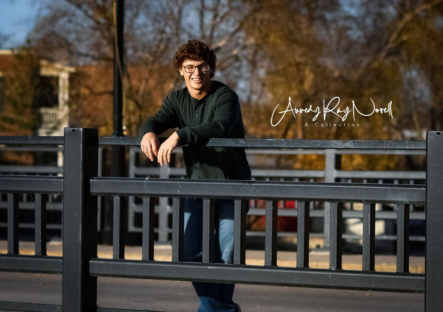 Casual outdoor portrait on wooden bridge railing with autumn trees in background.