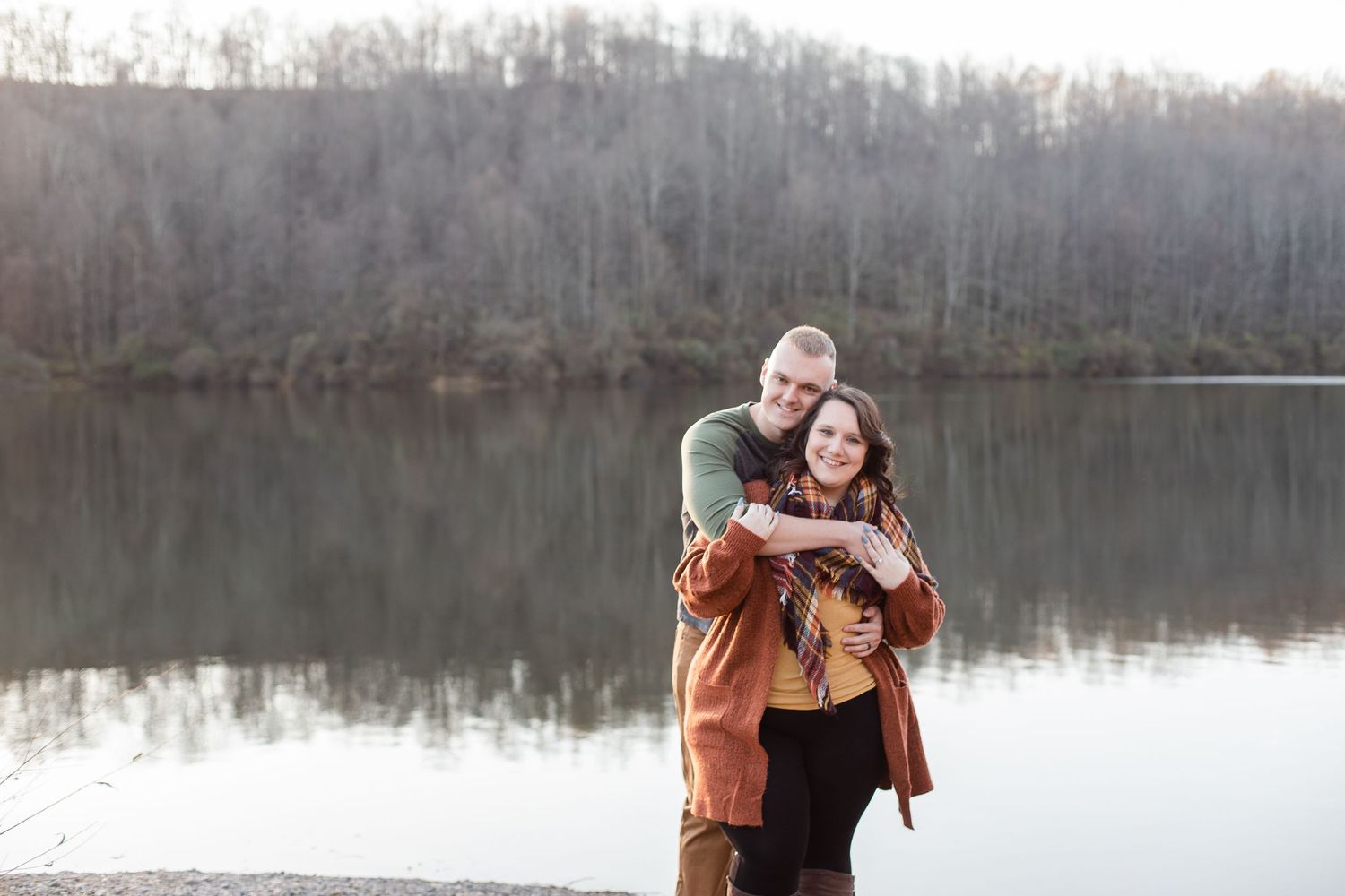 Couple shares affectionate moments by a peaceful lake with bare winter trees in the background.