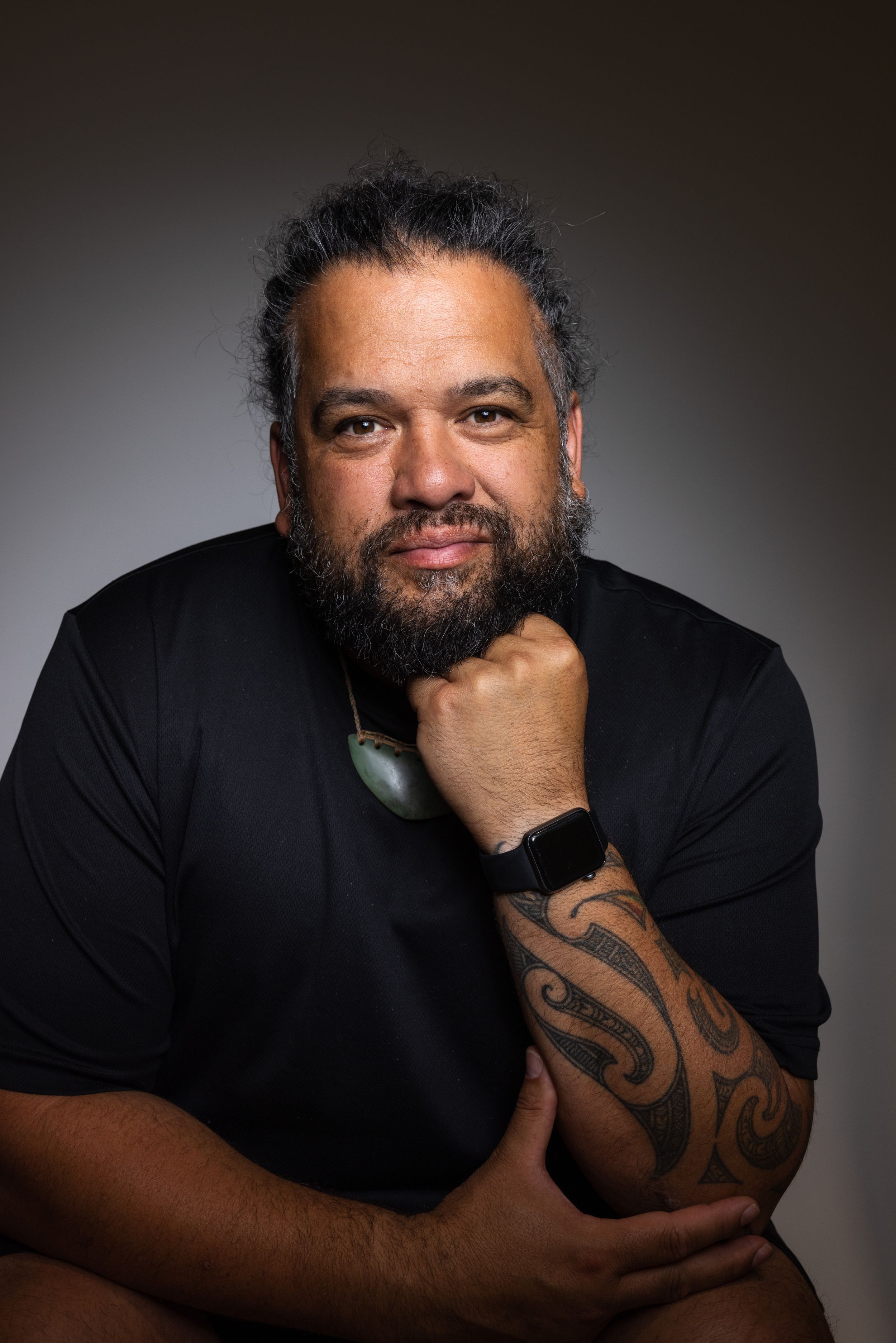 Studio portrait of person in black shirt with traditional tattoo and jewelry against dark background.