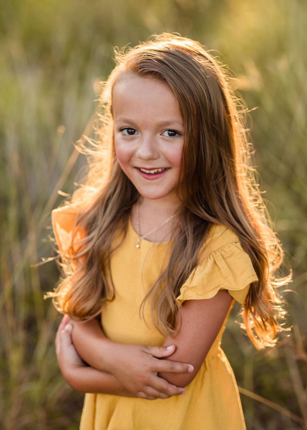 A child in a yellow dress poses playfully in a sunlit outdoor setting with tall grass and golden hour lighting.
