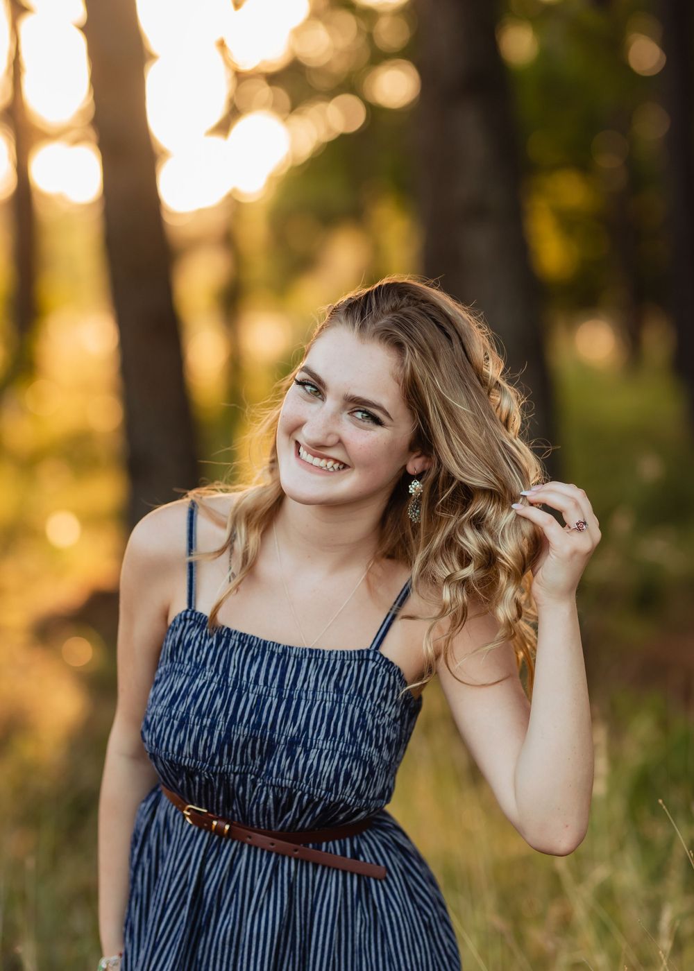 A senior graduate in a blue sundress with brown belt poses outdoors at golden hour with blurred bokeh background.