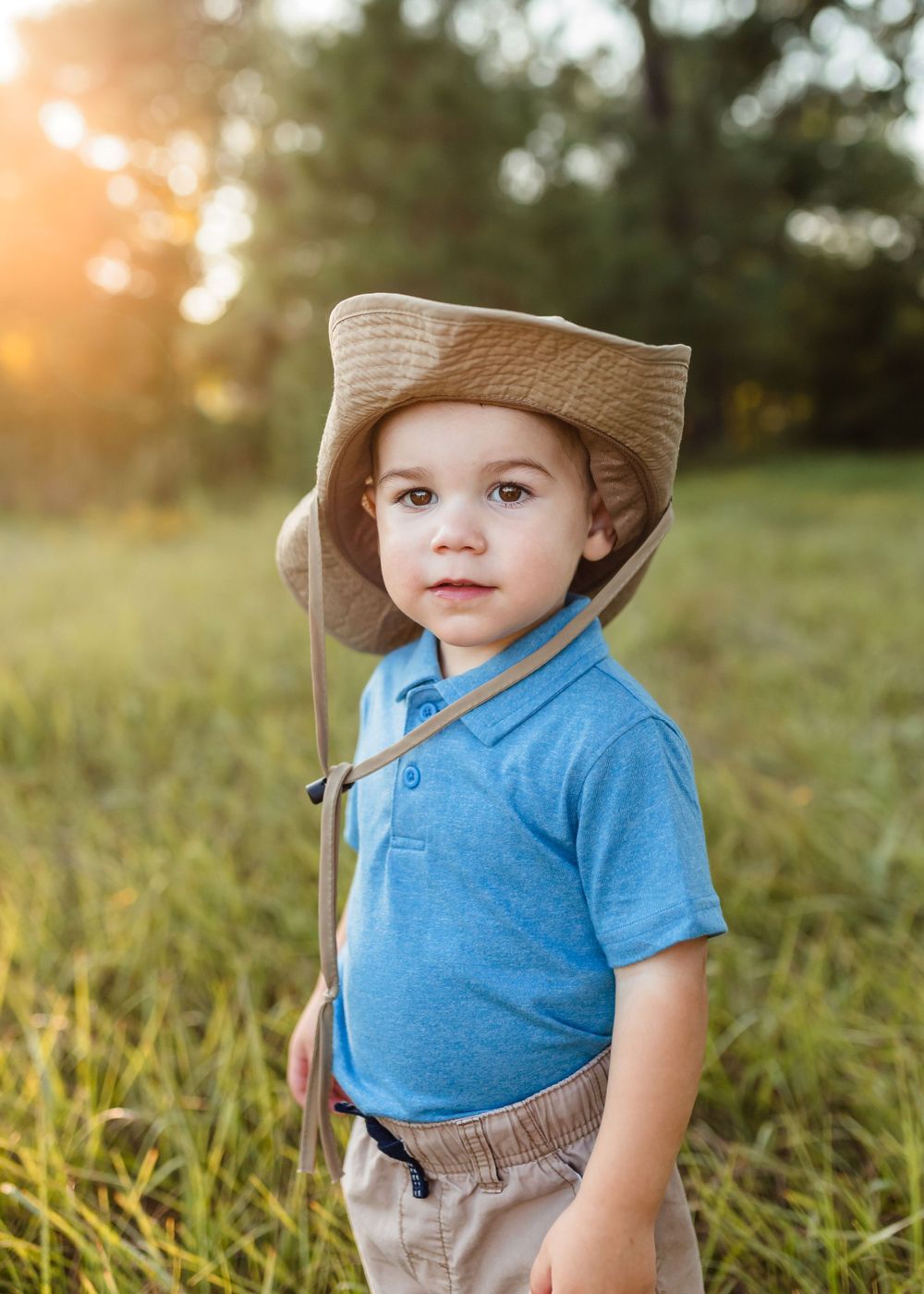 A toddler in a blue polo shirt and tan hat stands in a grassy field at sunset.