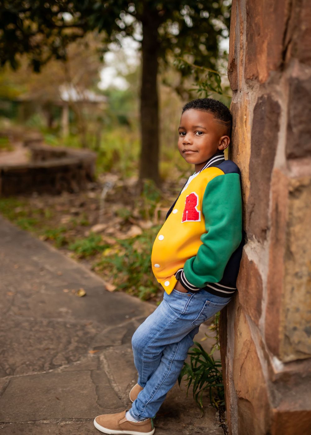 A young child in blue jeans and a green and yellow top leans playfully against a rustic post outdoors.