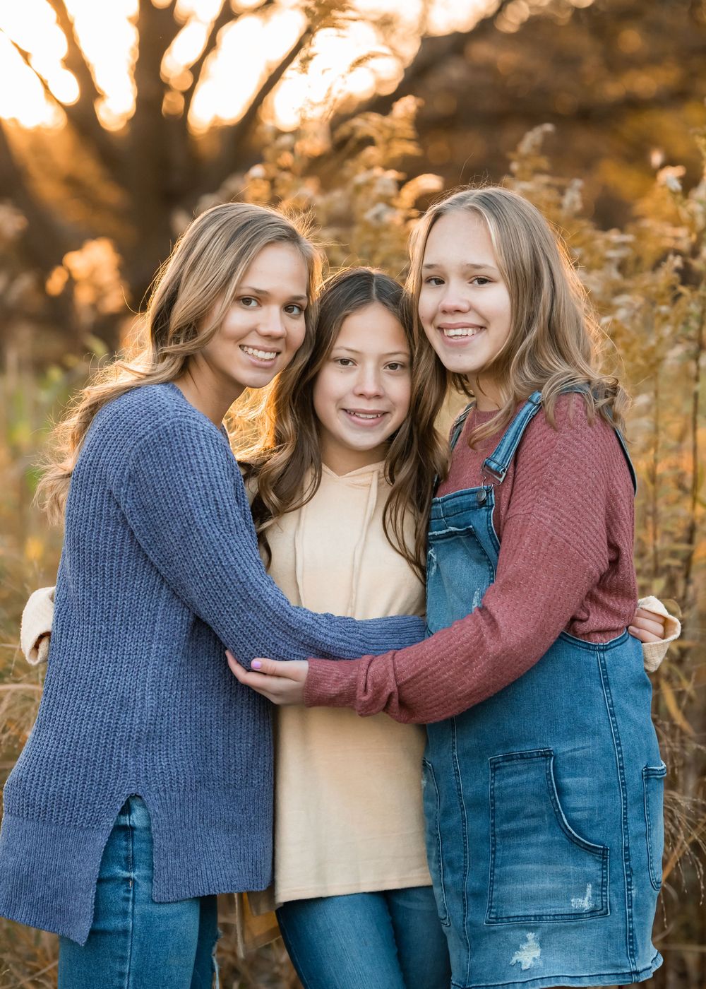 Three sisters in casual attire share warm embraces while posing together during a golden autumn photoshoot.