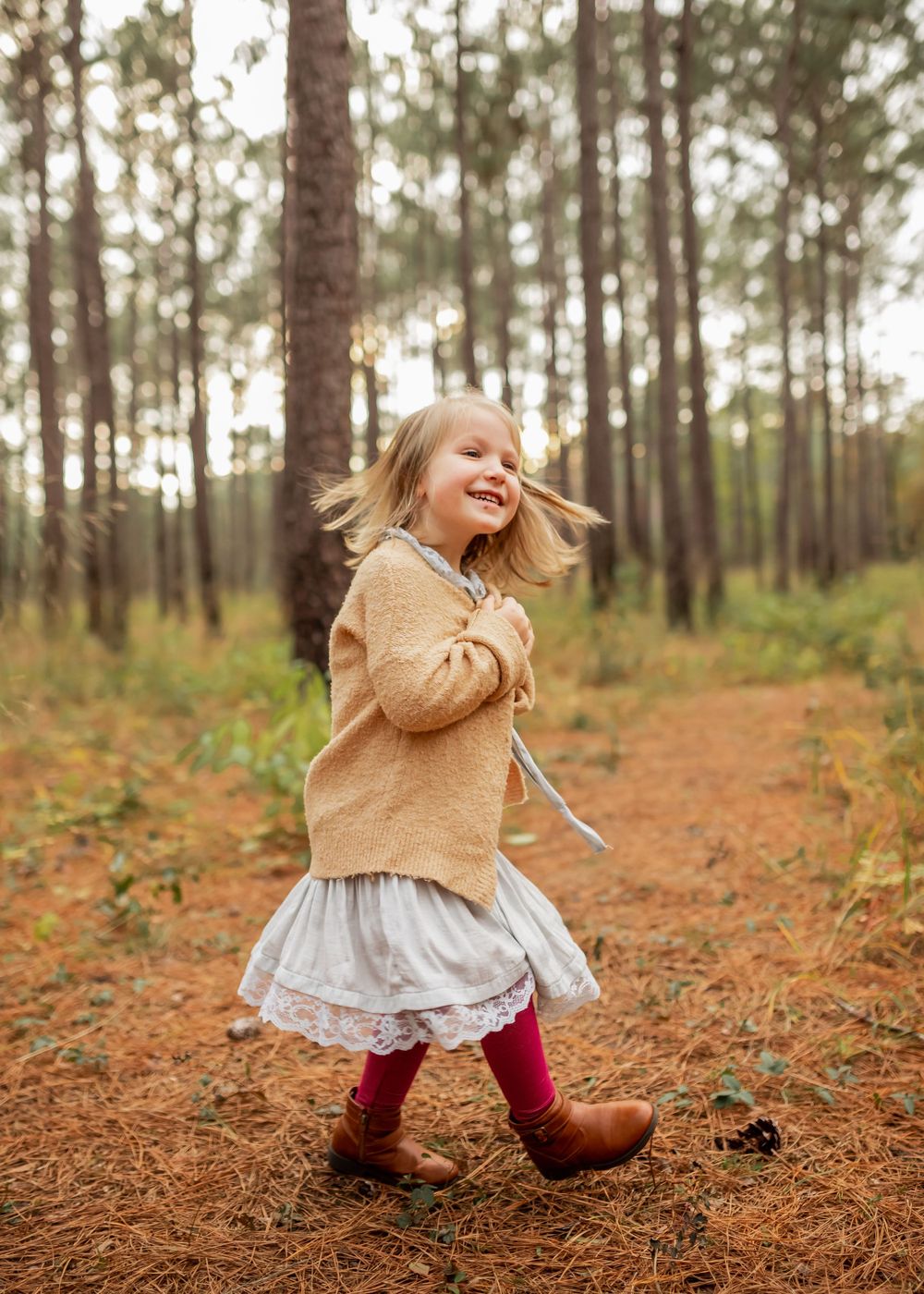 A young child in a brown sweater and dress skips playfully through a pine forest covered in fallen needles.