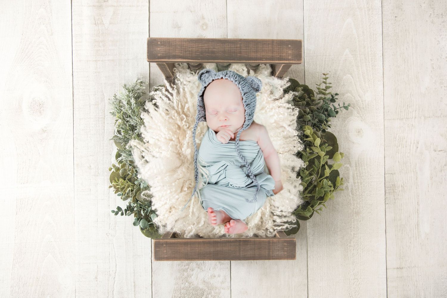 Newborn sleeping peacefully in wooden crate with white fluff and greenery decorations.