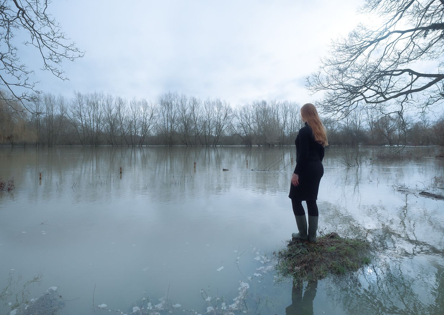 a woman in black stands looking away on a small island of grass in a flooded river