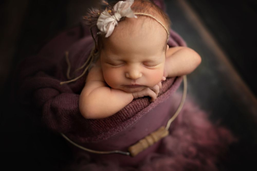 Sleeping newborn wearing floral headband nestled in purple wrap against dark background.