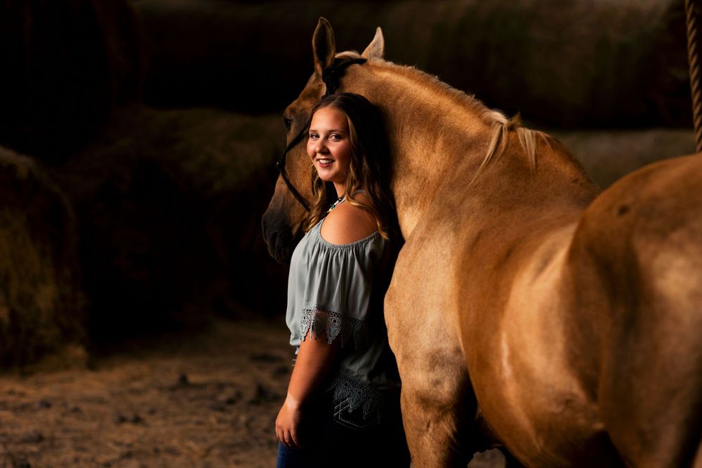 Young woman in grey dress standing next to beautiful brown horse at sunset.