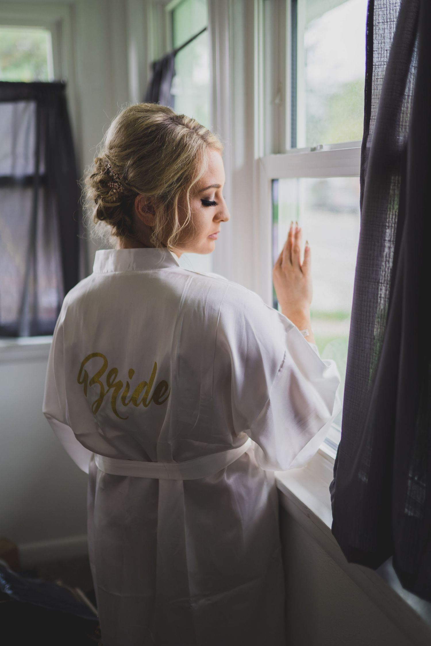 Person in white robe with 'Bride' text looking out window in soft morning light.