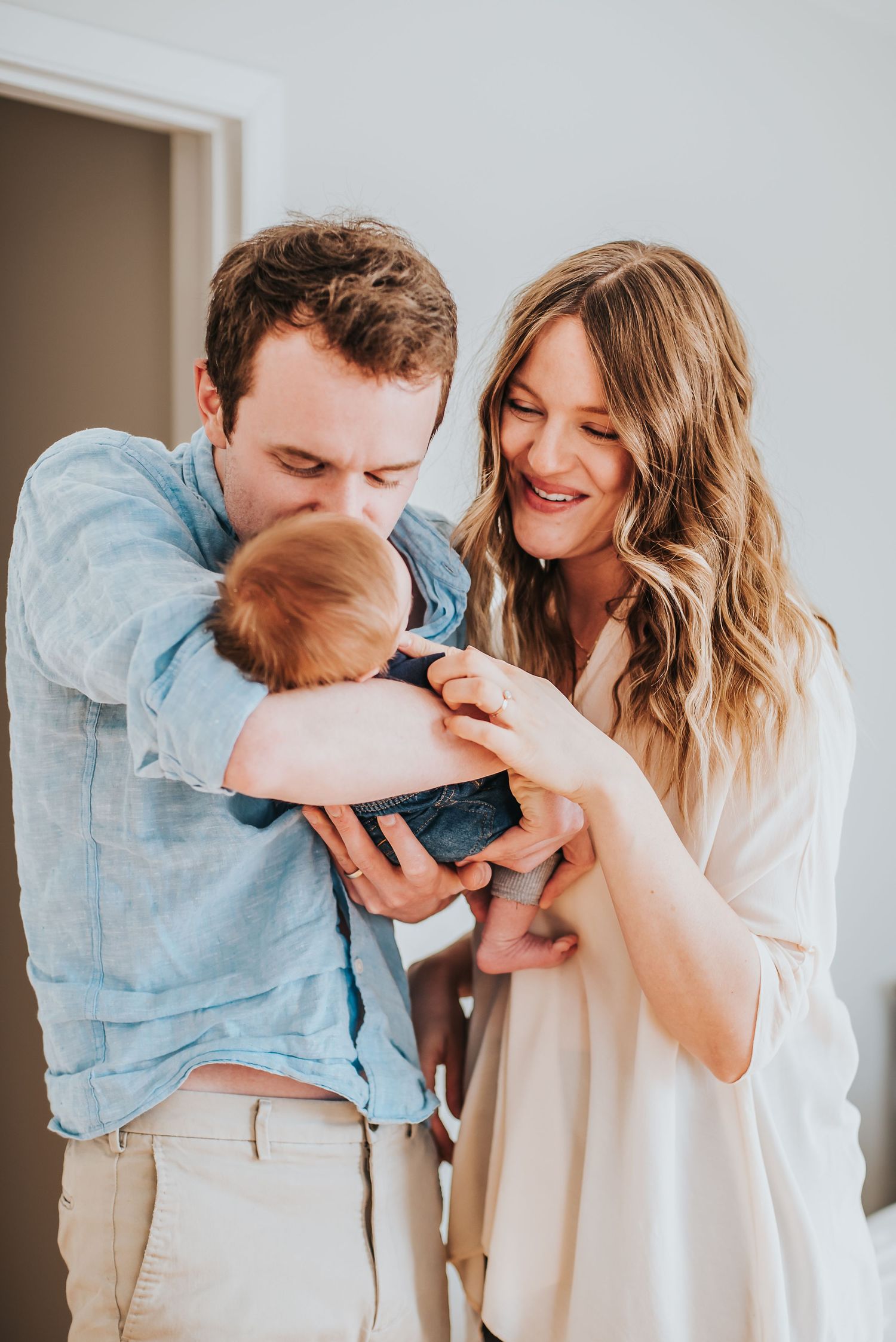 Caucasian man in blue shirt holds and kisses his newborn as his wife smiles.