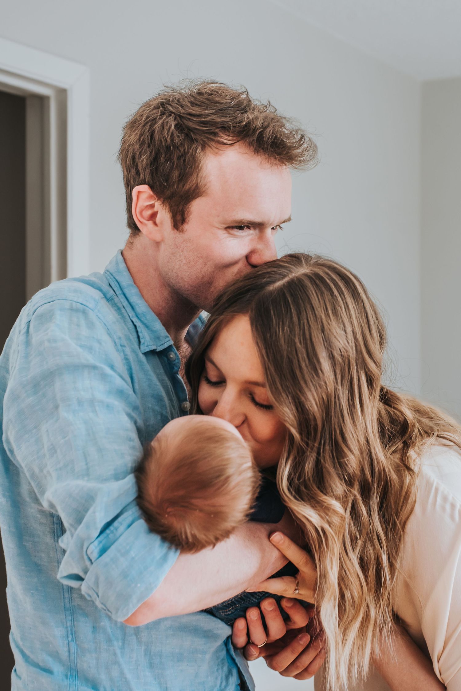 Caucasian man and woman hold one another and their newborn child, the man is kissing the woman's head as she kisses baby