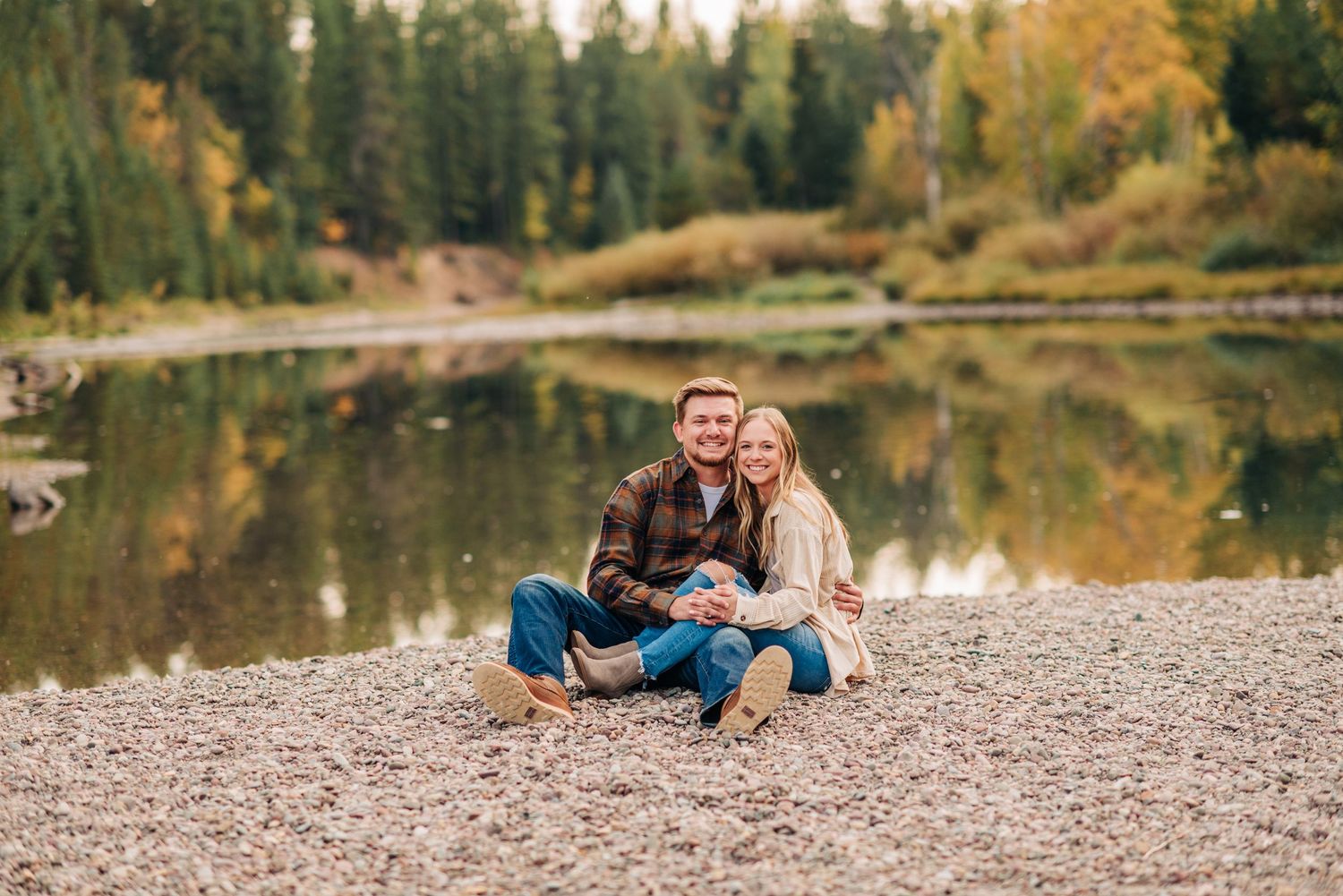 Couple sitting together on a rocky beach with autumn forest and reflective lake in the background.