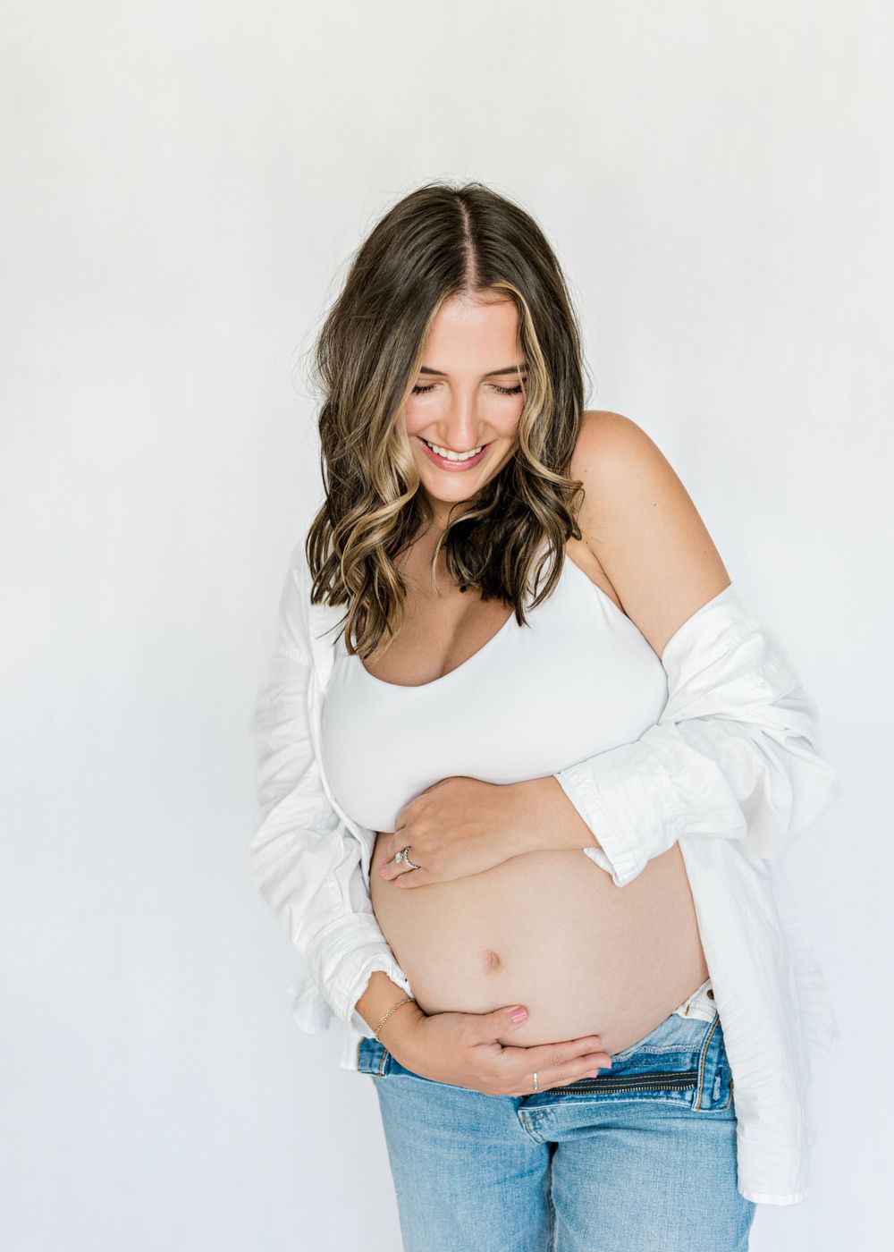 An expectant mother in white top and jeans smiles while cradling her baby bump.