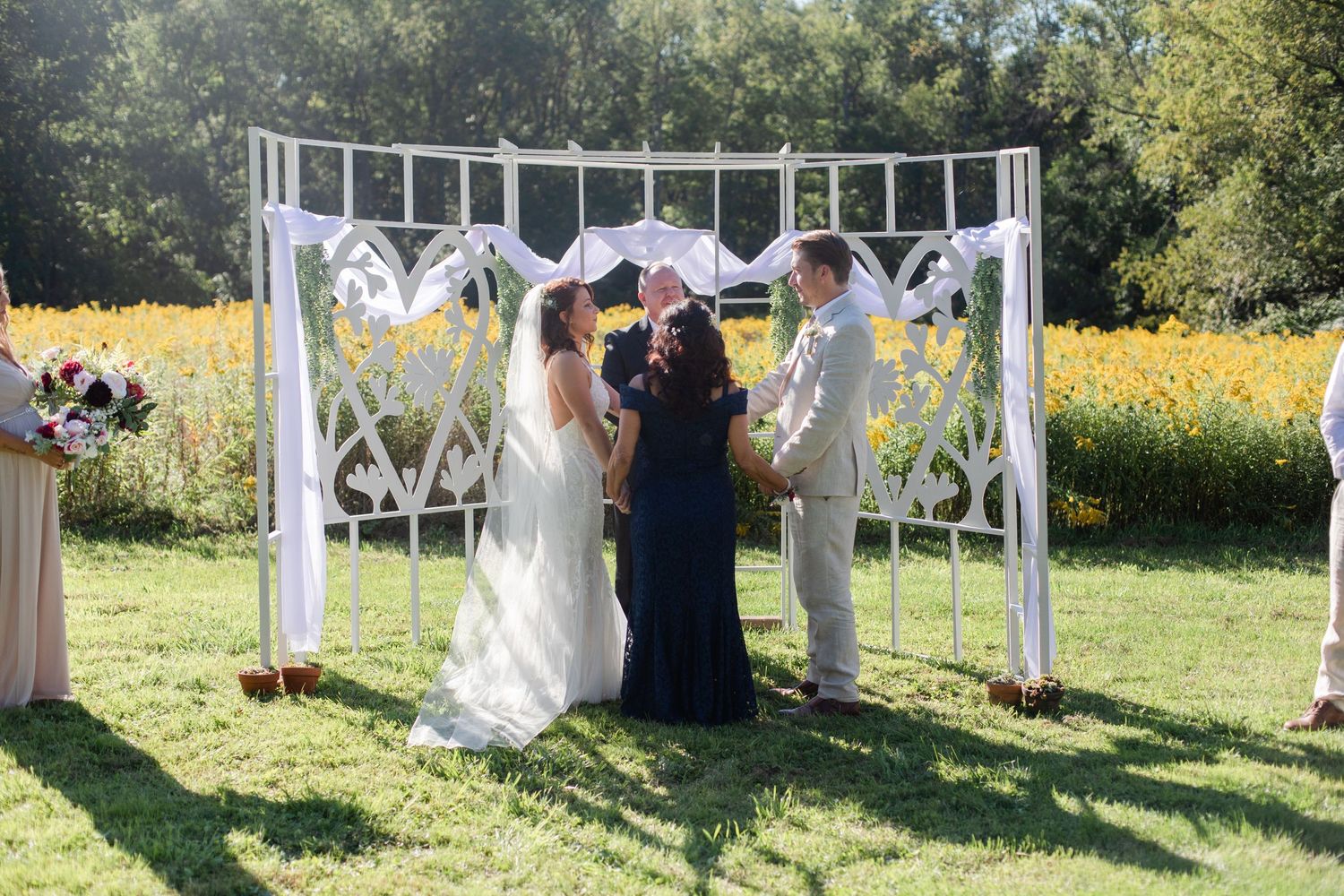 A wedding ceremony arch decorated with white fabric stands in a sunny field.