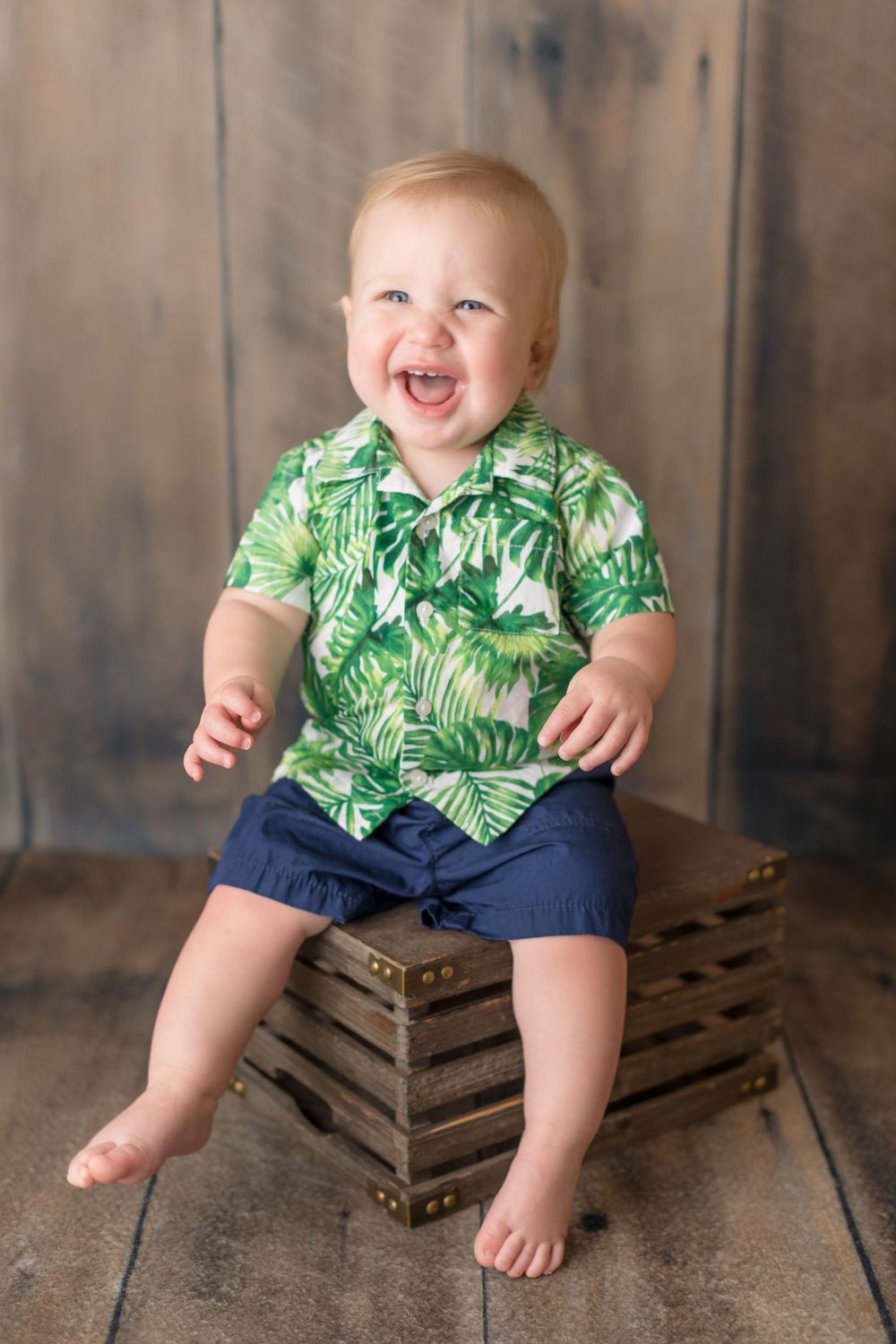 Toddler in tropical print shirt sitting on wooden crate against rustic background.