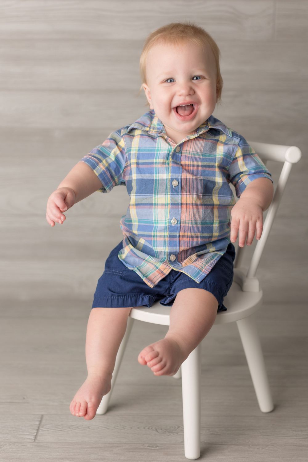 Happy baby wearing plaid shirt and navy shorts sits on white chair with joyful expression.